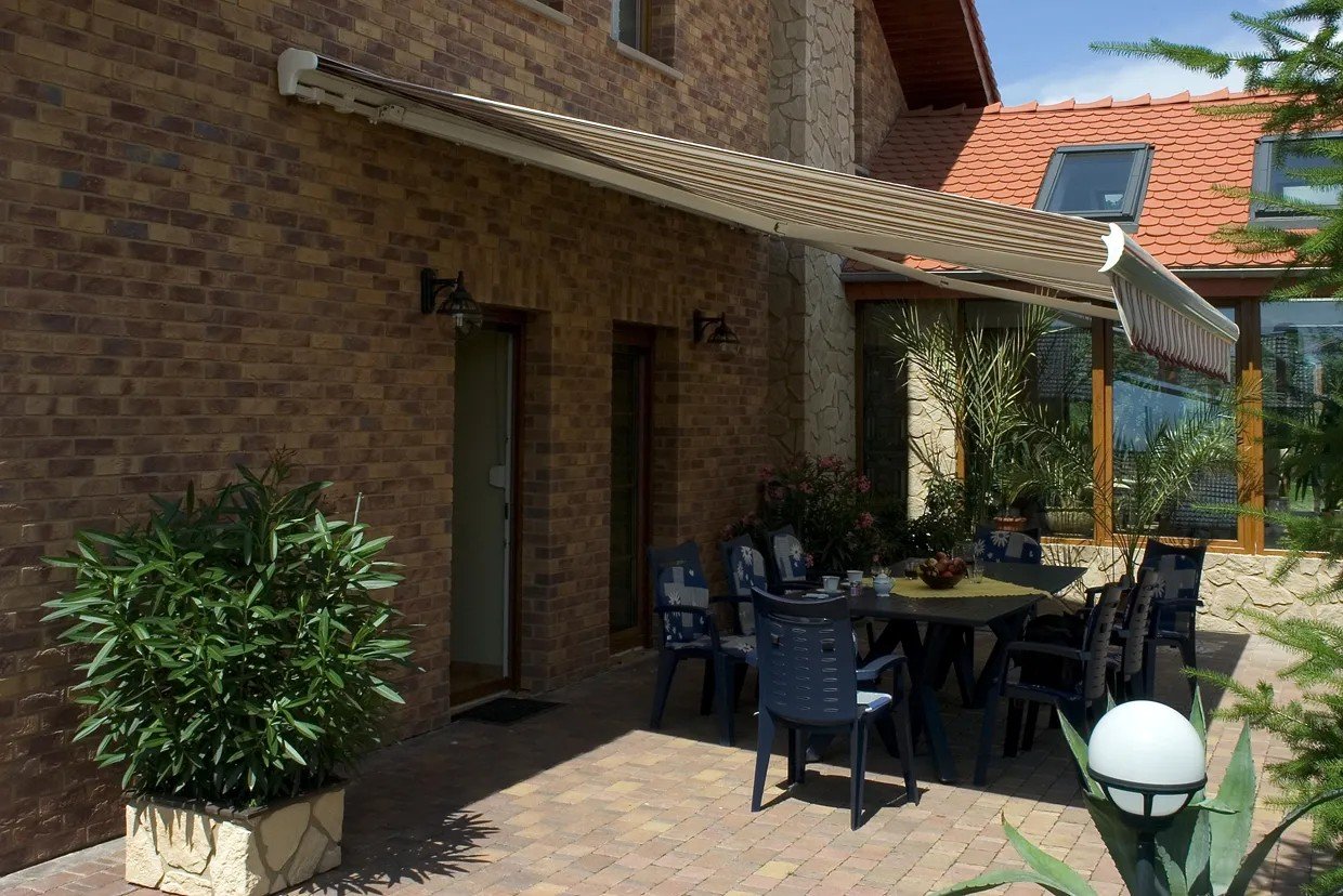 Uteområde med bord og stoler under en utendørs markise, omgitt av planter og en murvegg, med en del av huset i bakgrunnen.