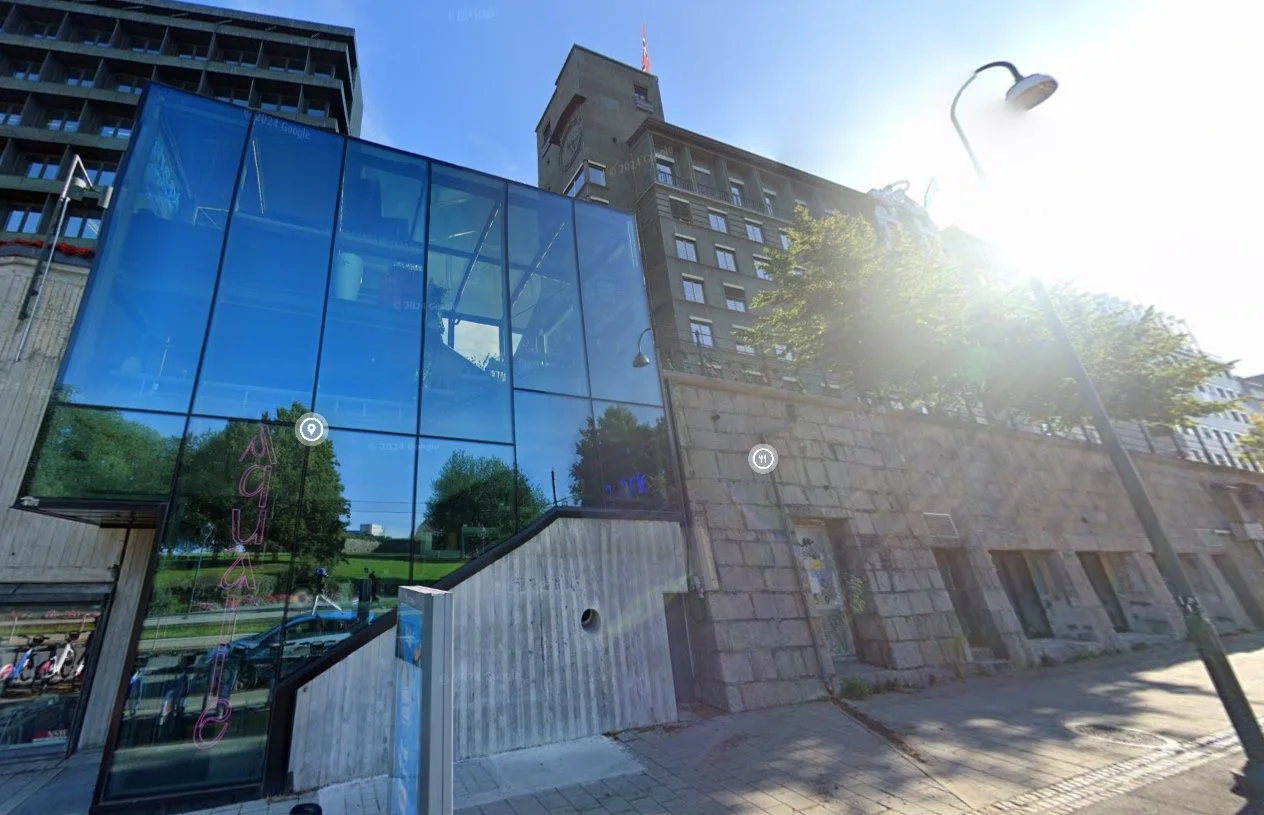 Bygning med glassfasade og en høy konsentrasjon av vinduer, omgitt av trær og gatebelysning, med en blå himmel og solskinn.