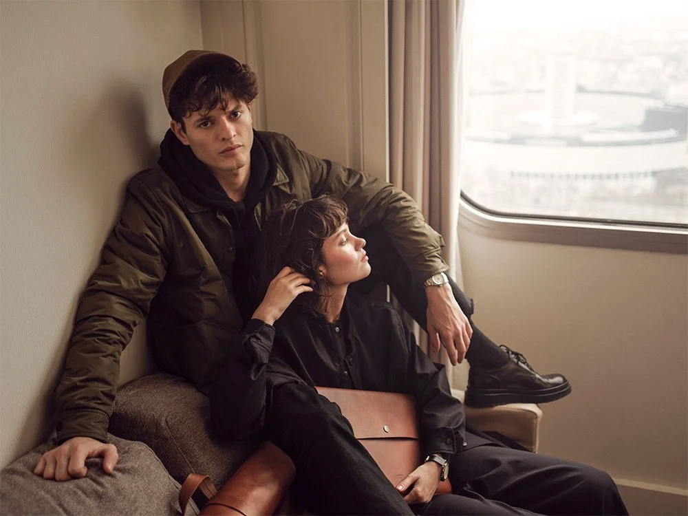 Two people sitting on a couch near a window, one wearing a brown jacket and cap, the other dressed in black, holding a brown leather bag.