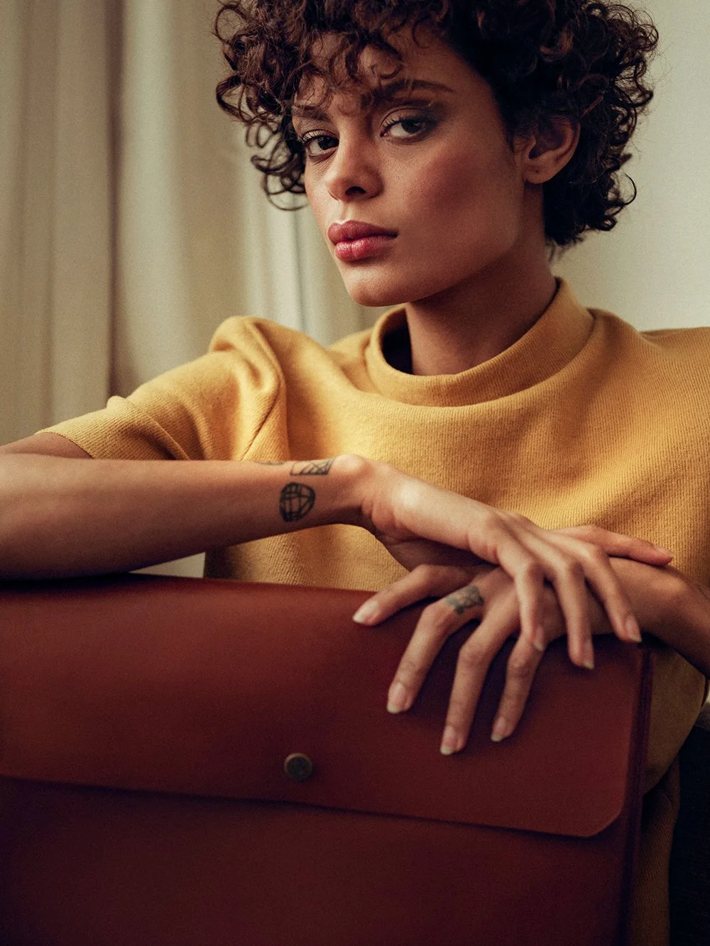 Fashion model in yellow top holding a brown leather bag, showing tattoos on fingers.
