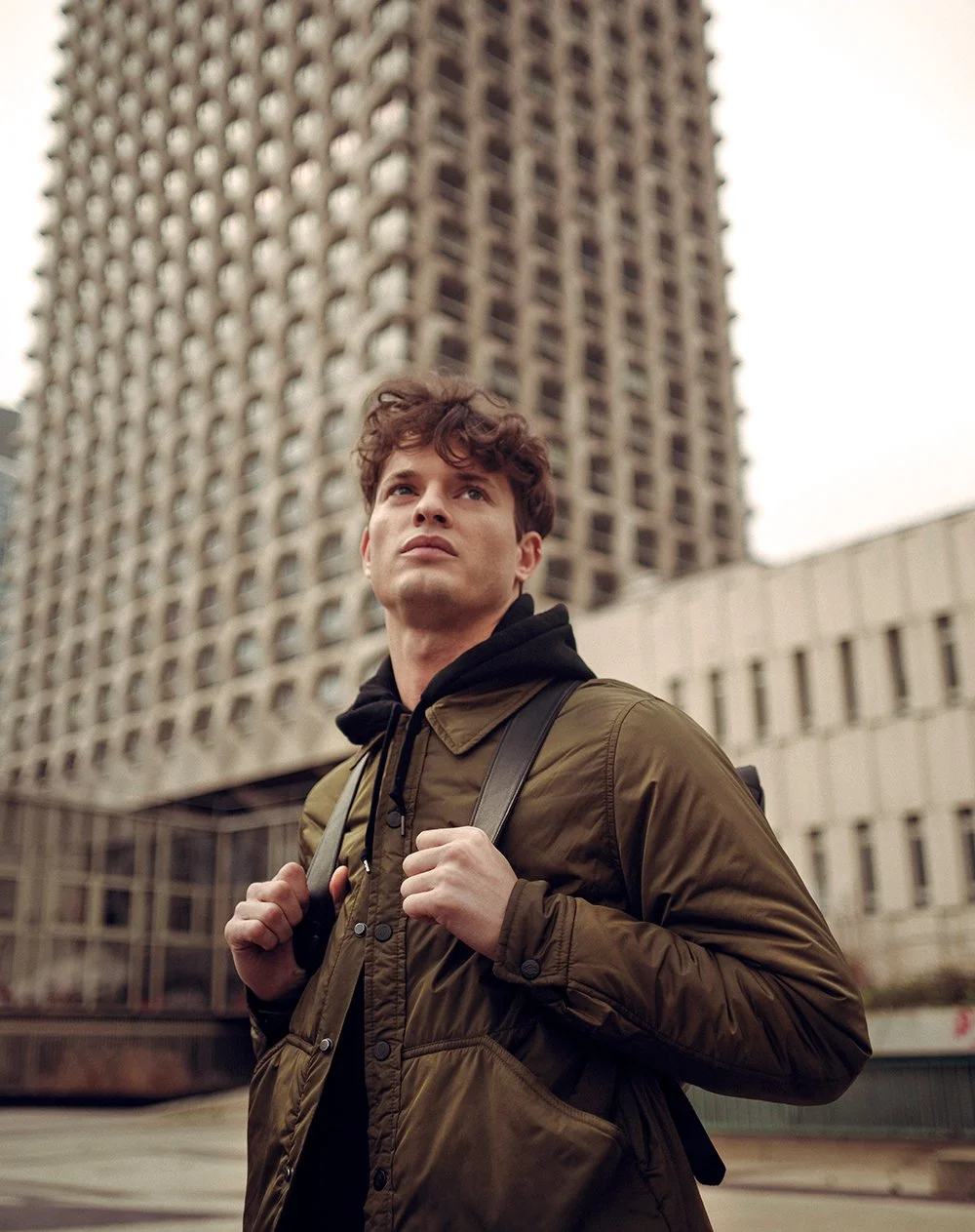 Man in green jacket with backpack standing in front of tall building, looking upwards.