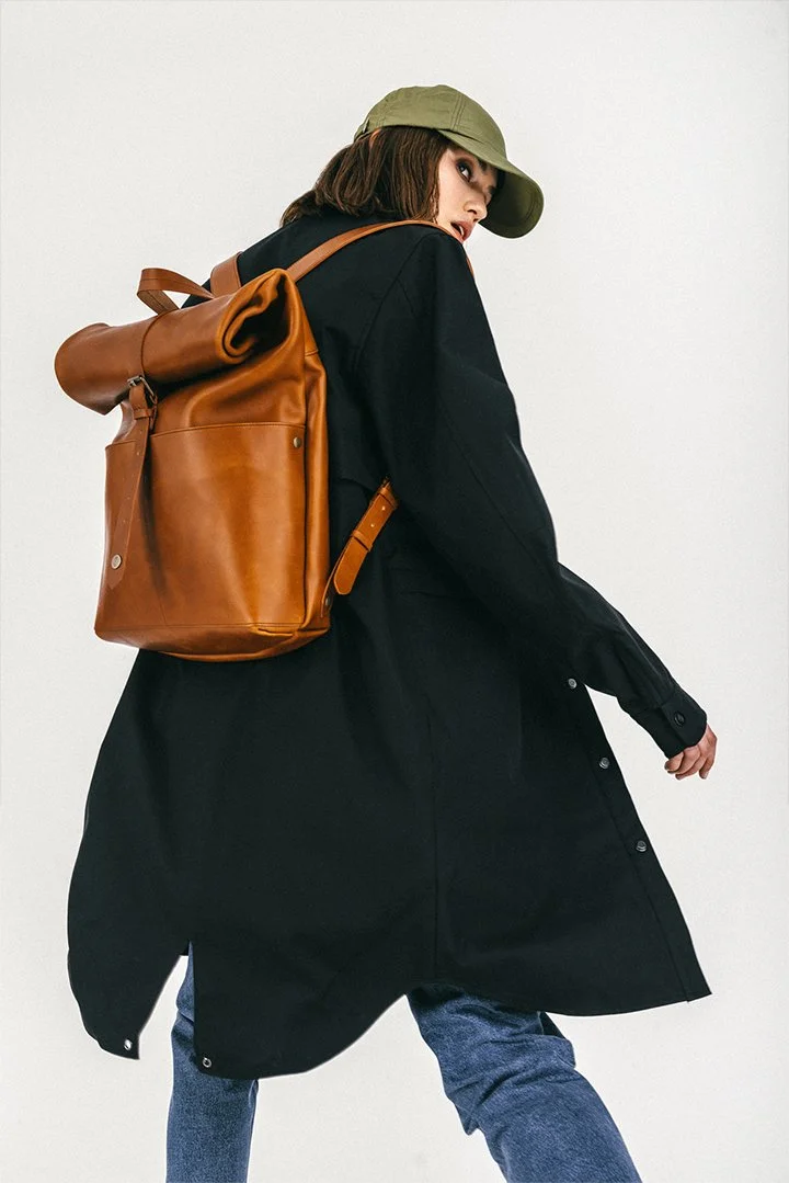 Person wearing black coat, jeans, green cap, and brown leather backpack