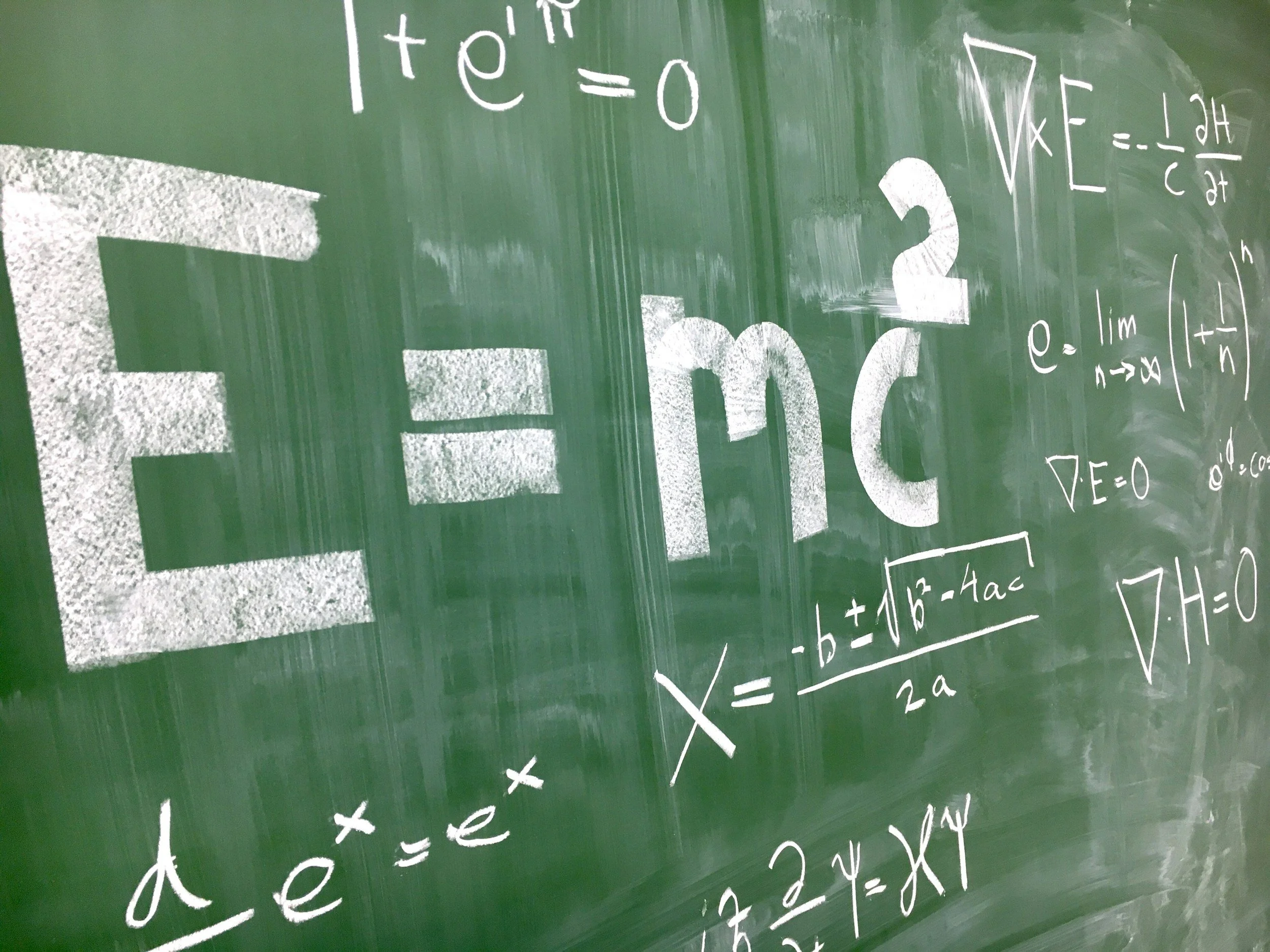 A green chalkboard covered with various mathematical equations and formulas written in white chalk, including the famous equation E=mc^2 and quadratic formula.