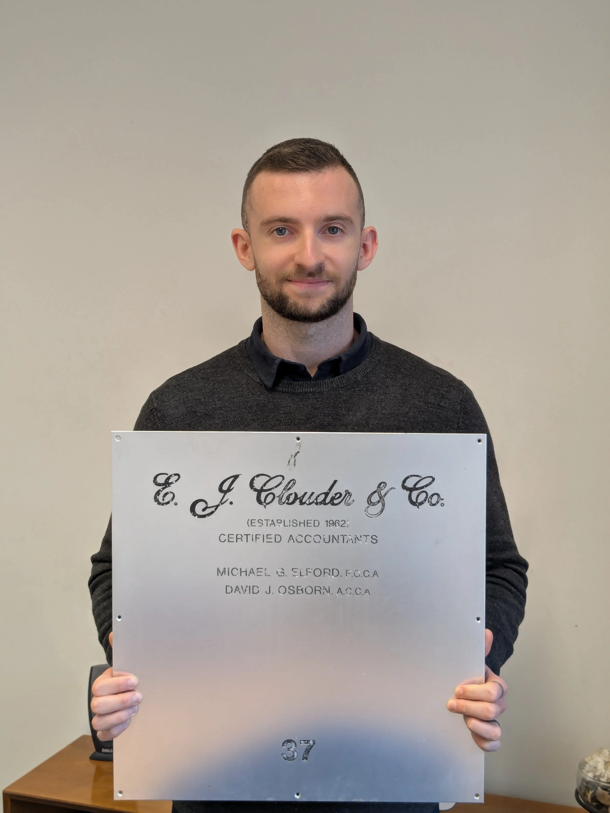 A young man with short brown hair and a beard, dressed in a dark gray sweater over a collared shirt, holding a silver plaque with black text. The plaque reads 'E. J. Clouder & Co.', established 1962, Certified Accountants, Michael G. Elford, David J. Osborn, and the number 37. He is standing in front of a plain beige wall.