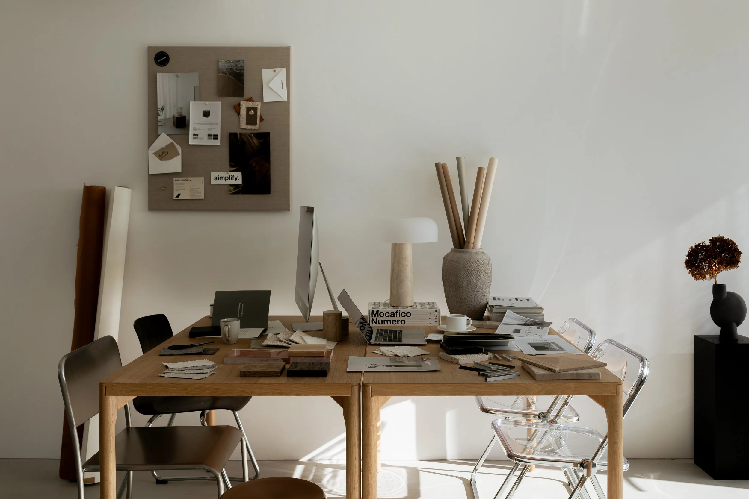 Modern, minimalist interior design studio with wood desks, lucite chairs, an inspiration board on the wall and fabric and material samples on the desk