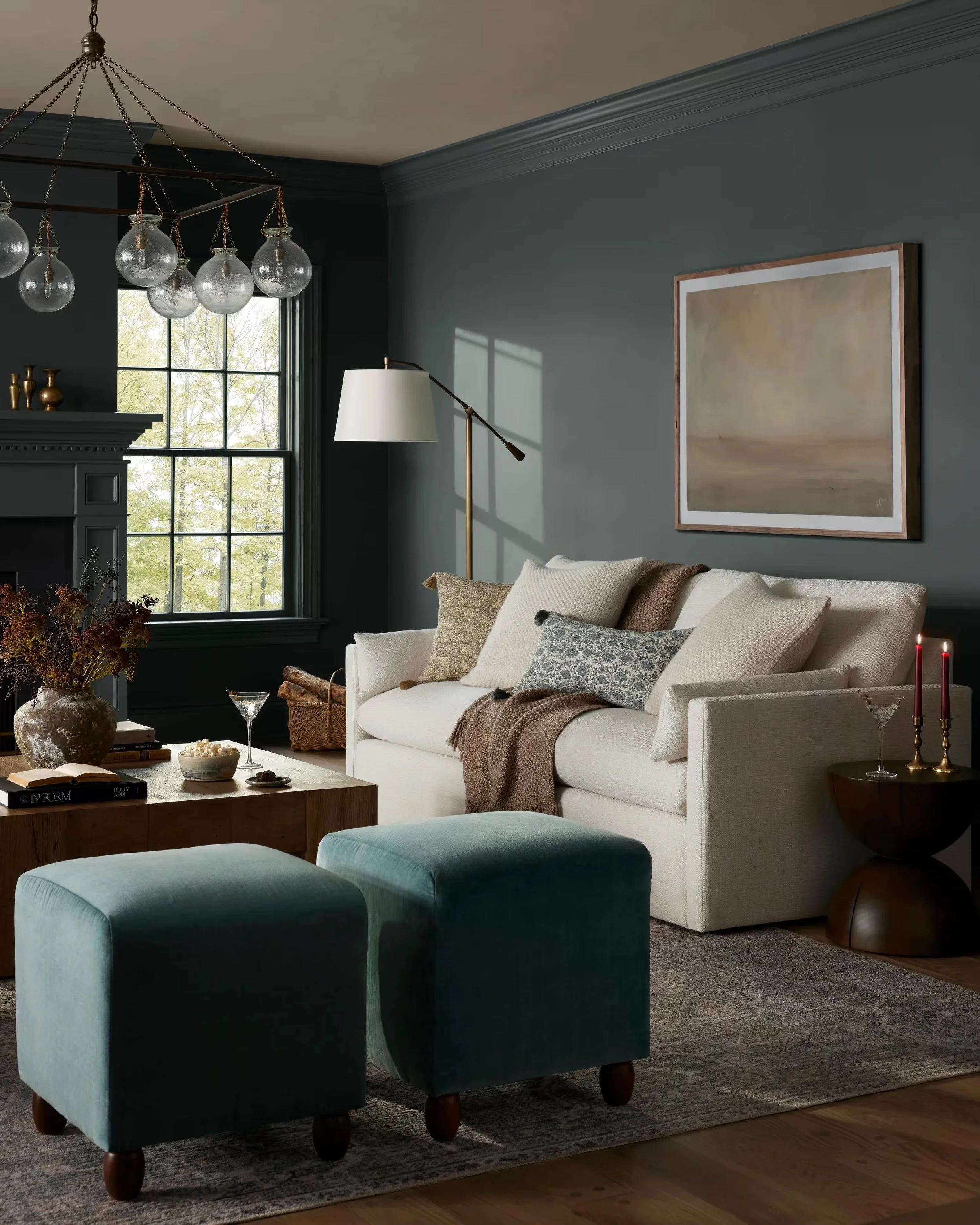 A living room with a moody feel - rich blue painted walls with matching velvet ottomans, a neutral oat color sofa with layered pillows and throws in similar and contrasting colors, large wood coffee table in front of an ornate millwork fireplace.