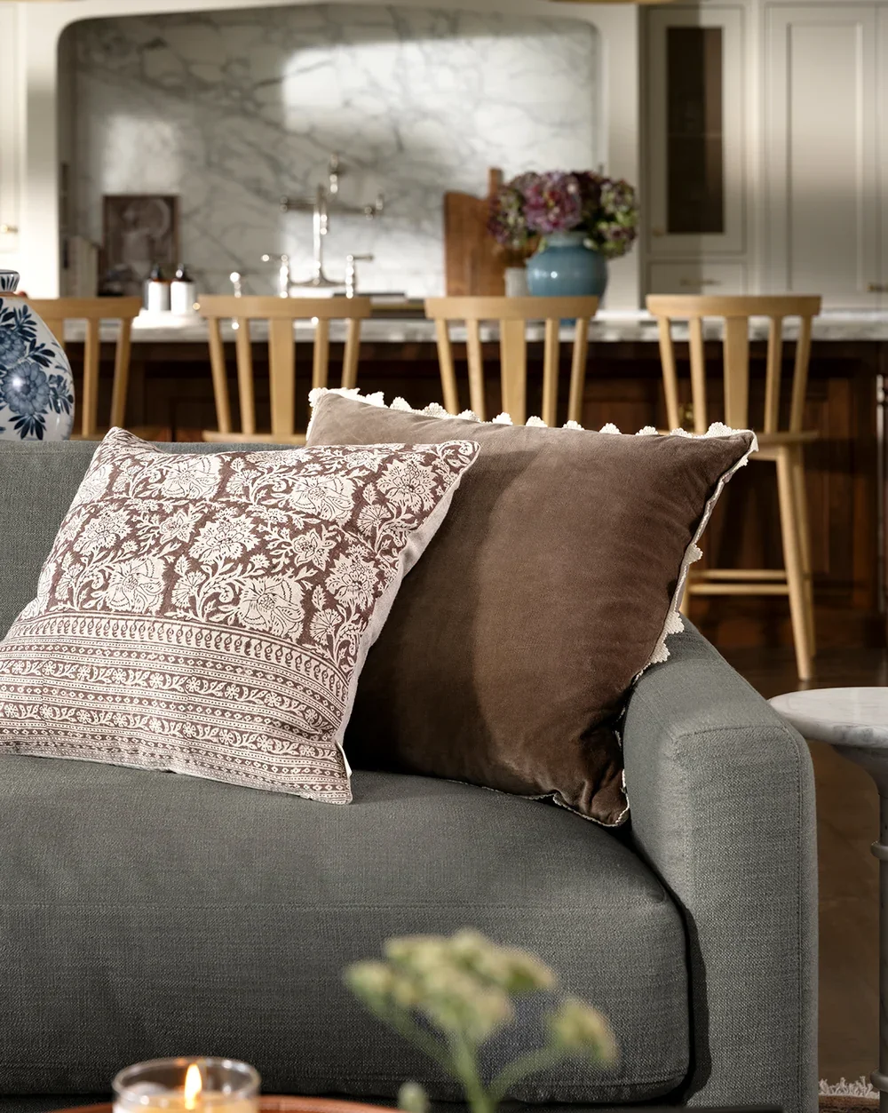 Combination of scallop edge detail velvet pillow and floral and stripe print fabric pillow on a grey sofa with a farmhouse modern style kitchen in the background