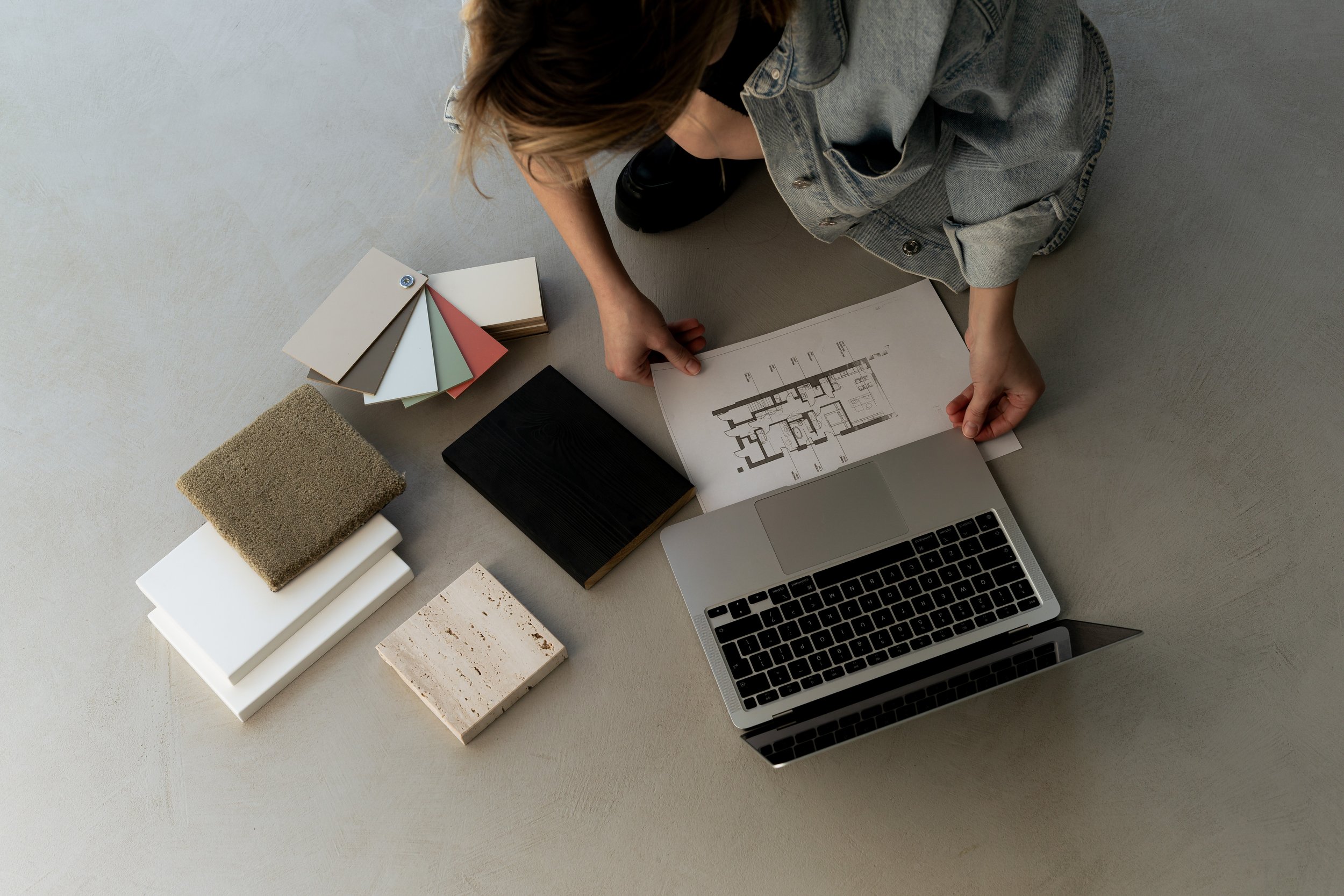Minimalist photoshoot with a woman sitting on the floor in front of a laptop with fabric and paint samples and a floor plan.