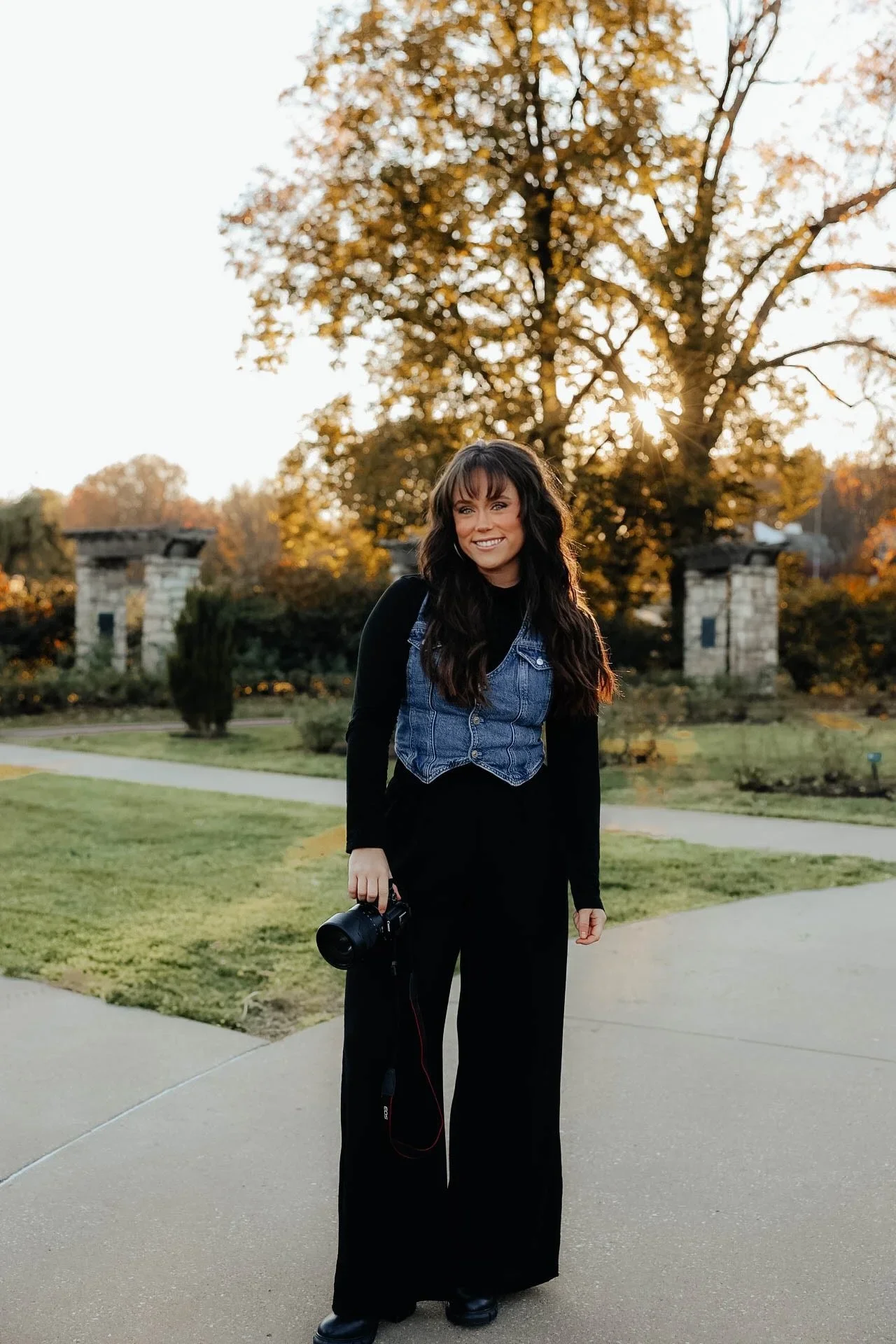 A woman with long dark curly hair smiling and holding a camera, standing outdoors on a sidewalk with trees and a decorative stone gate in the background during sunset.