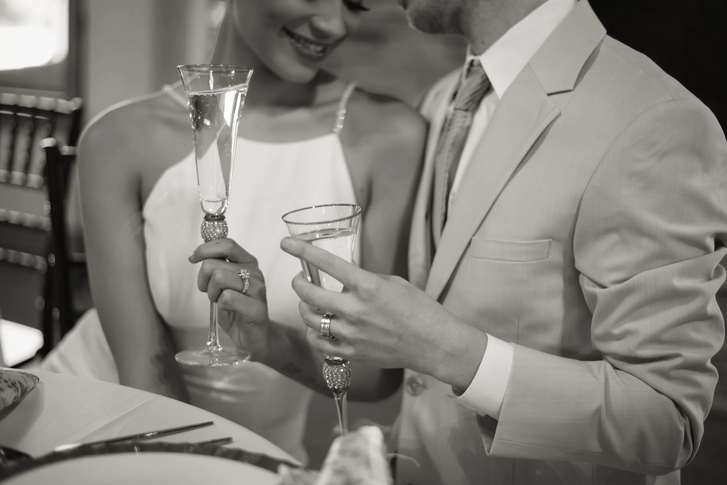 A couple dressed in formal attire holding champagne glasses at a celebration or wedding, smiling and leaning close to each other.