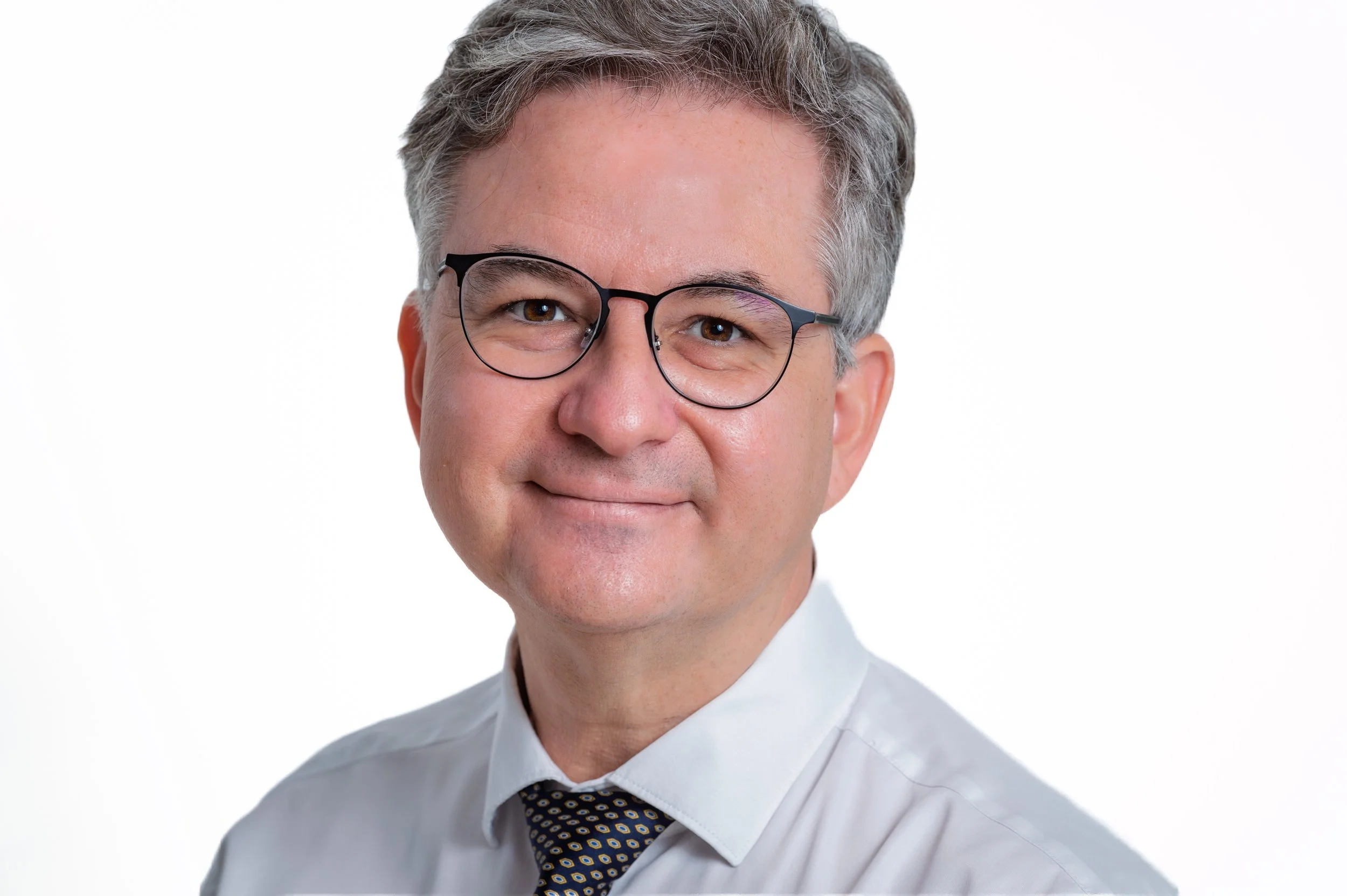 A professional portrait of Dr. Bernd Schmid, glasses, wearing a white shirt and patterned tie, smiling softly against a white background.