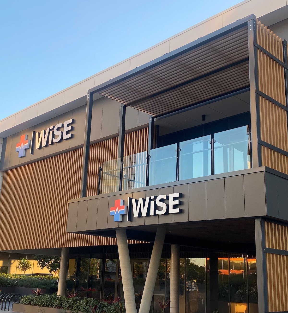 Modern multi-story building with the word 'WISE' and a medical cross logo on the facade, featuring glass balconies and wooden accents.
