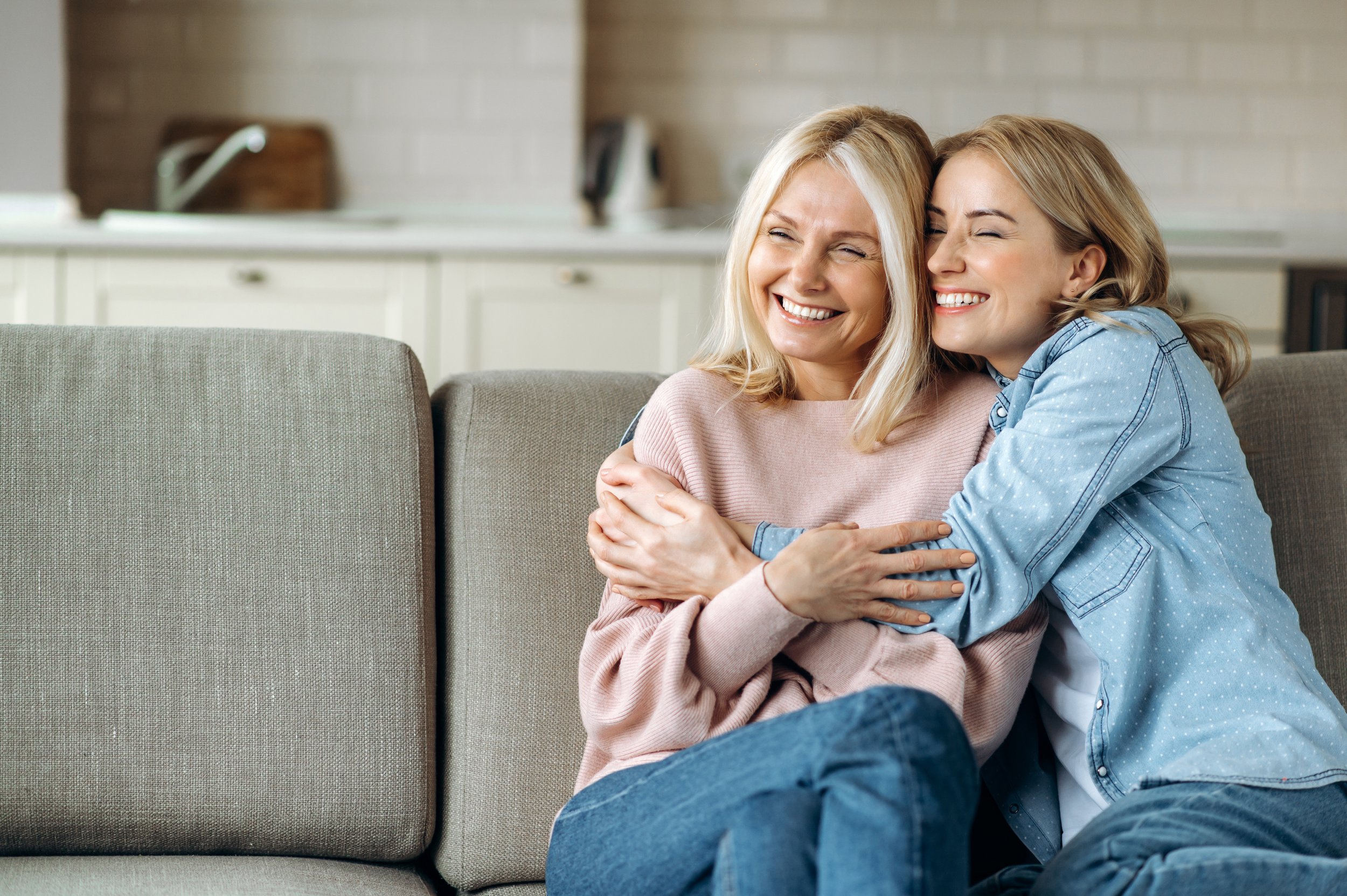 Mature attractive mother and her young adult happy daughter, are sitting at home on the couch, embracing, smiling.