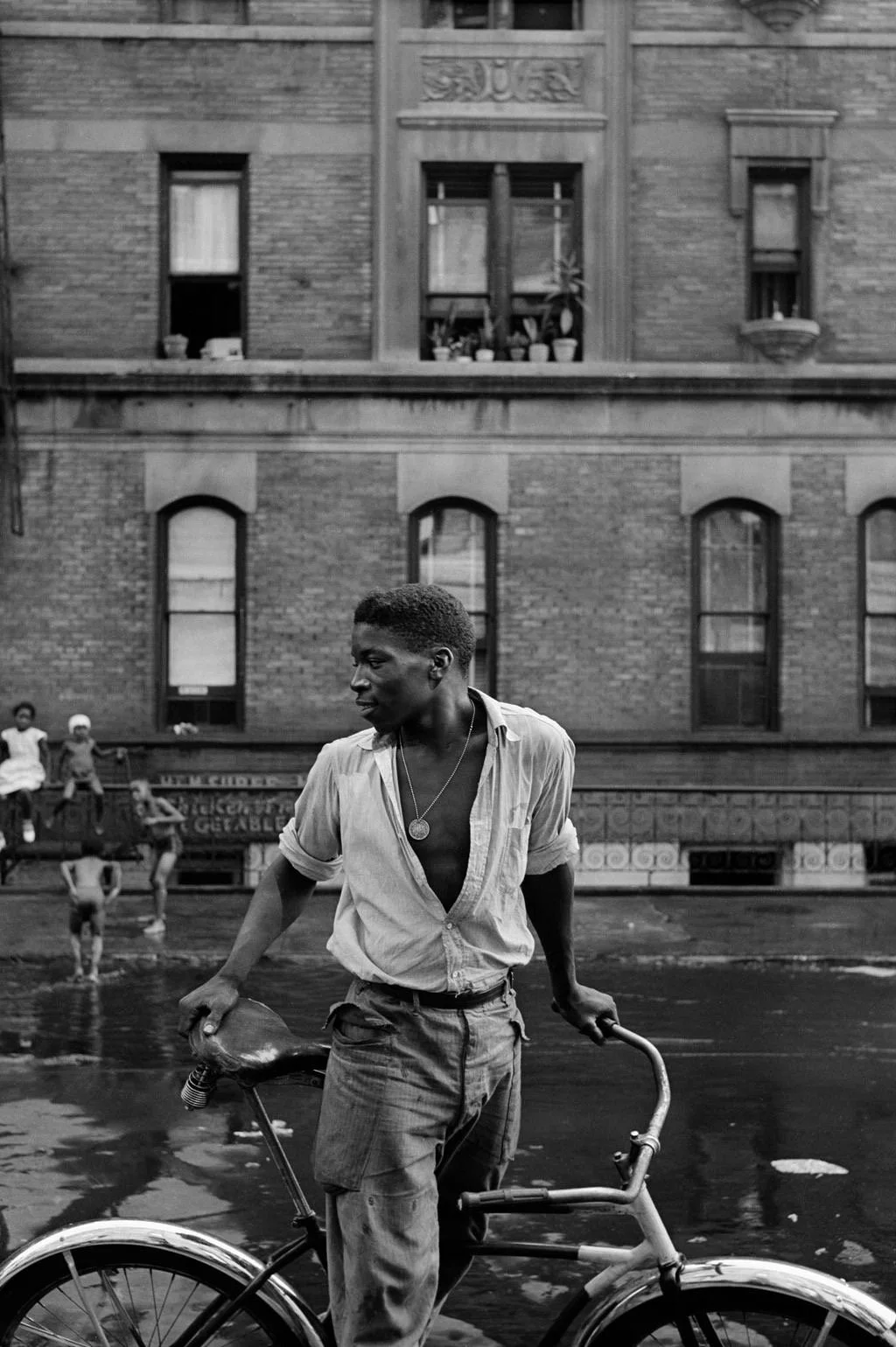 A young man with dark skin, wearing a partially unbuttoned shirt and pants, stands next to a bicycle on a city street. In the background, there are old brick buildings with multiple windows, some with plants and decorations, and children playing on a bench.
