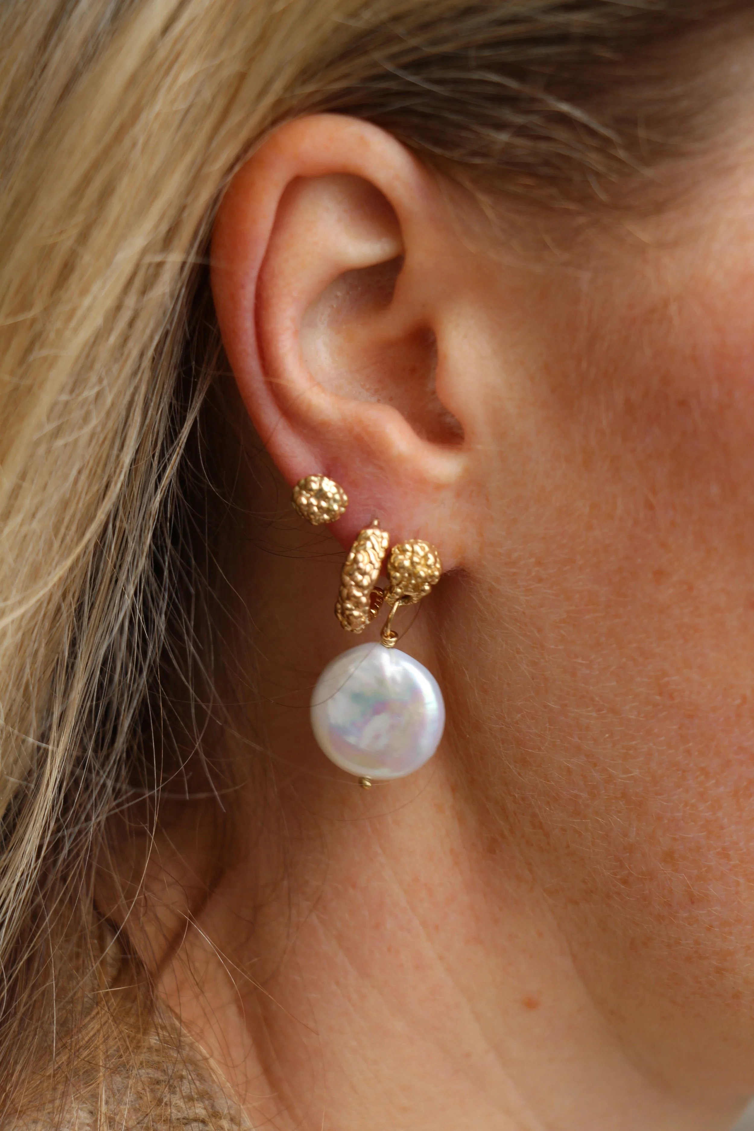 Close-up of a woman's ear with gold earrings and a large pearl drop earring, with blonde hair and skin visible.