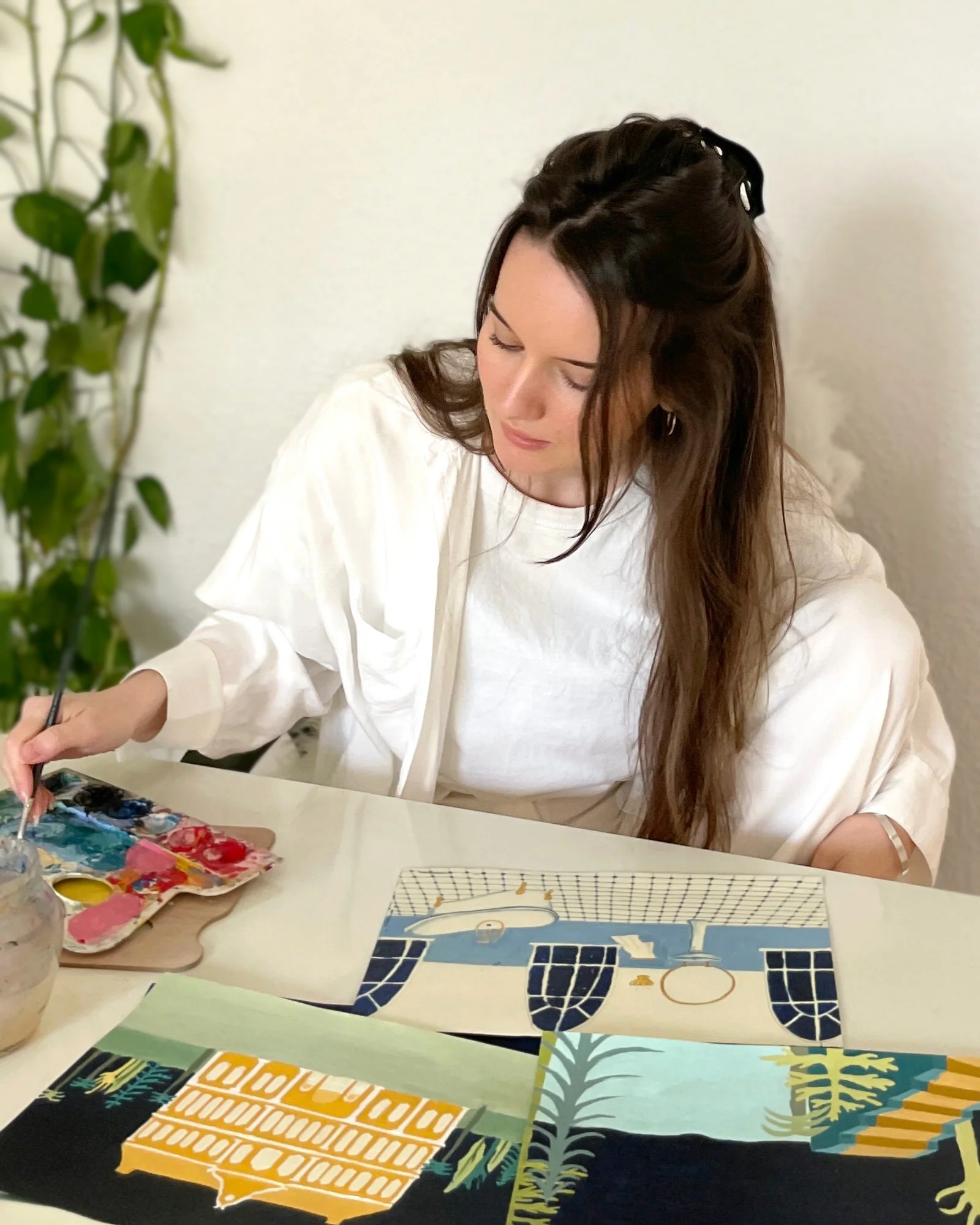 A woman with long brown hair, wearing a white long-sleeve shirt, is sitting at a table painting on canvas with a palette ofColors. There are multiple organized canvases with different designs on the table.