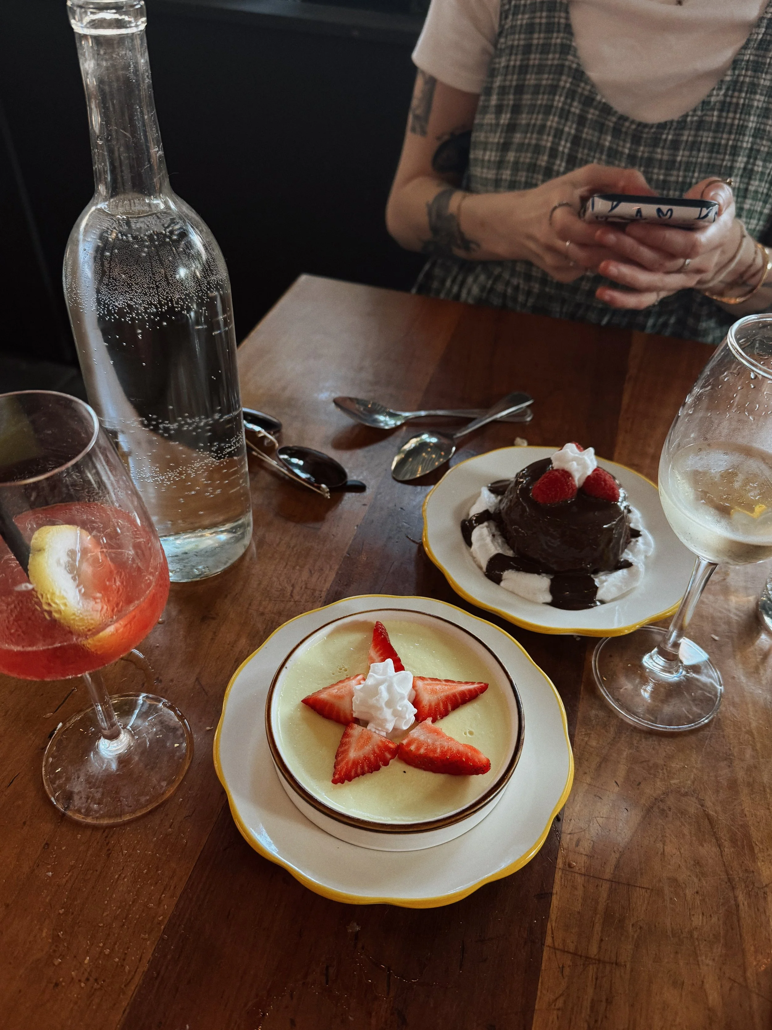 Table with three desserts: a strawberry tart, a chocolate cake with strawberries, and a cube-shaped cheesecake with strawberries, surrounded by drinks, cutlery, sunglasses, and a person using a phone.