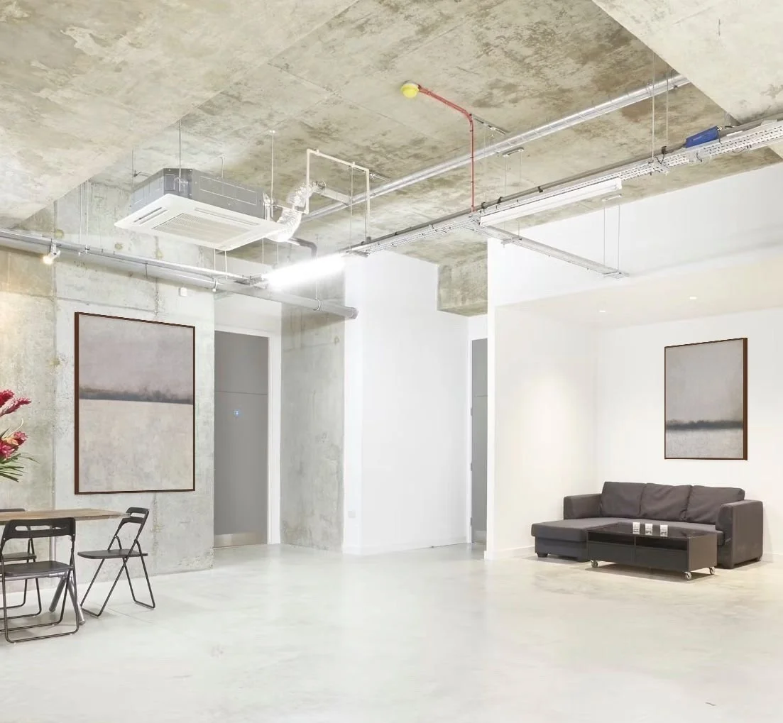 Modern, minimalist interior with concrete ceiling, white and light gray walls, a black sofa with a small table, framed artwork on the walls, a dining area with black chairs, and visible ceiling ductwork and electrical wiring.