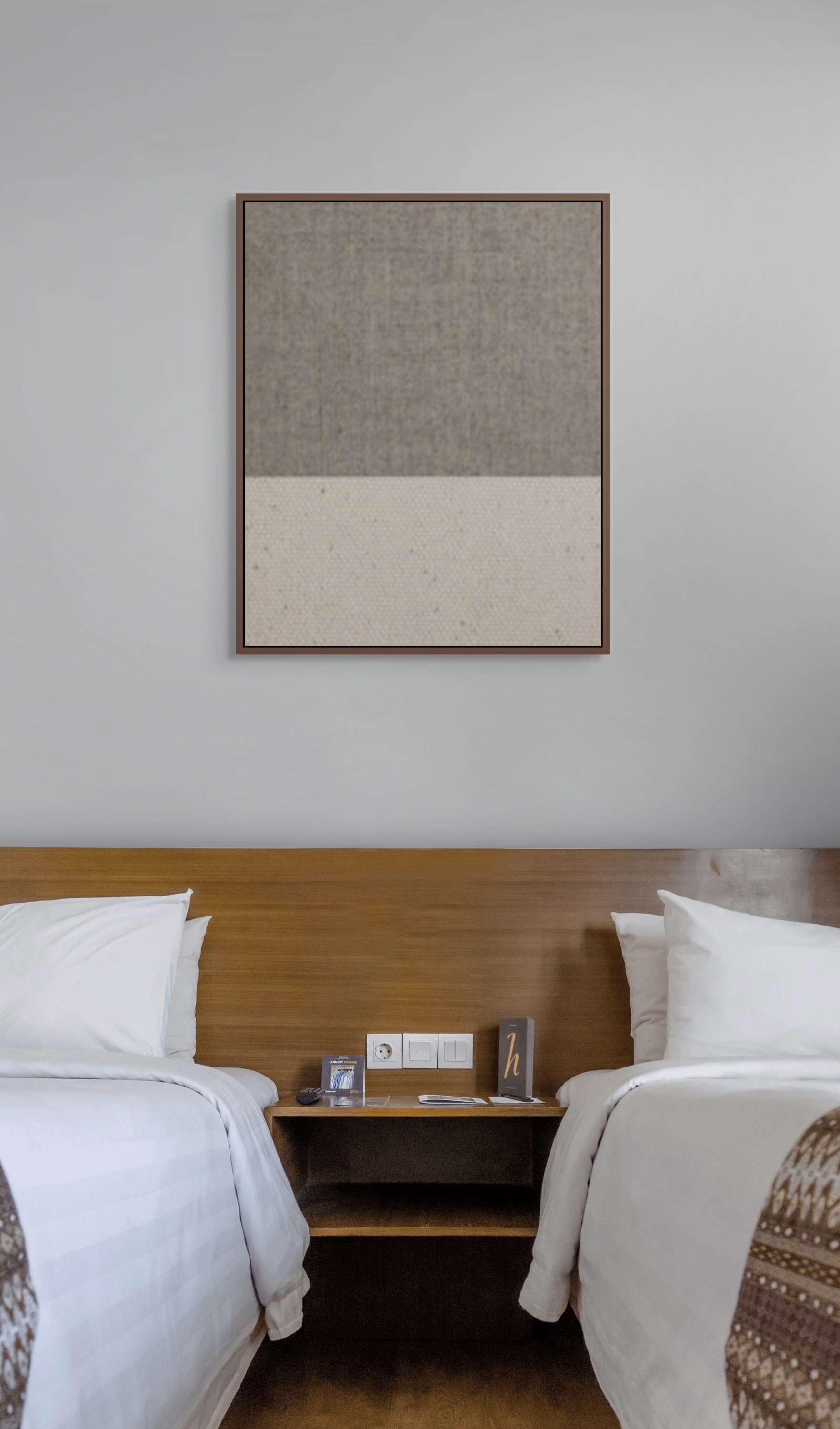 Two twin beds with white bedding and pillows, separated by a wooden nightstand, against a white wall with a large, framed textile art piece hanging above.