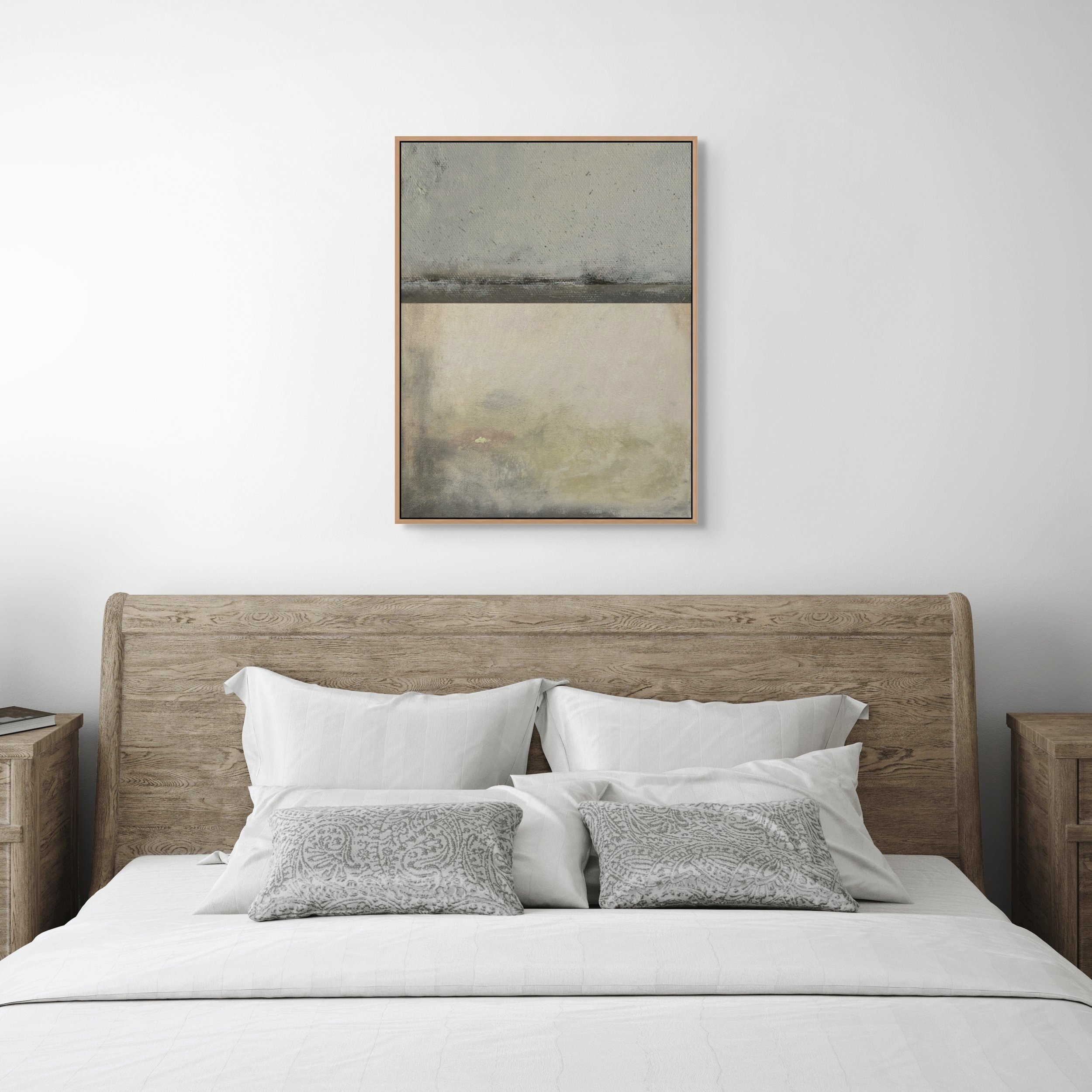 A neatly made bed with white pillows and patterned gray accent pillows, a wooden headboard, and matching wooden nightstands on each side. Above the bed is a framed abstract painting with muted tones, on a plain white wall.