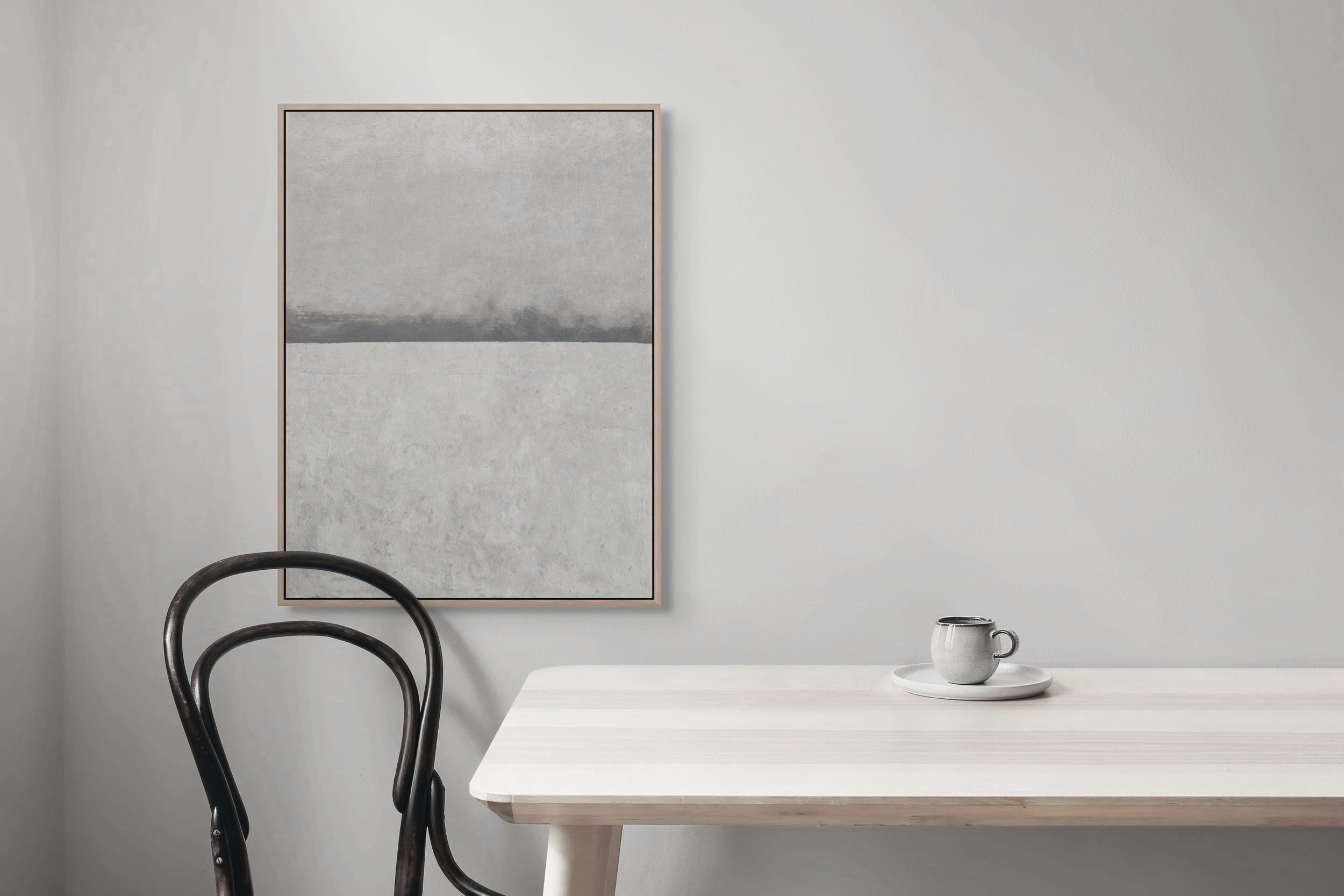 Minimalist dining area with a wooden table, a grey cup on a saucer, a black bentwood chair, and an abstract painting on a white wall.