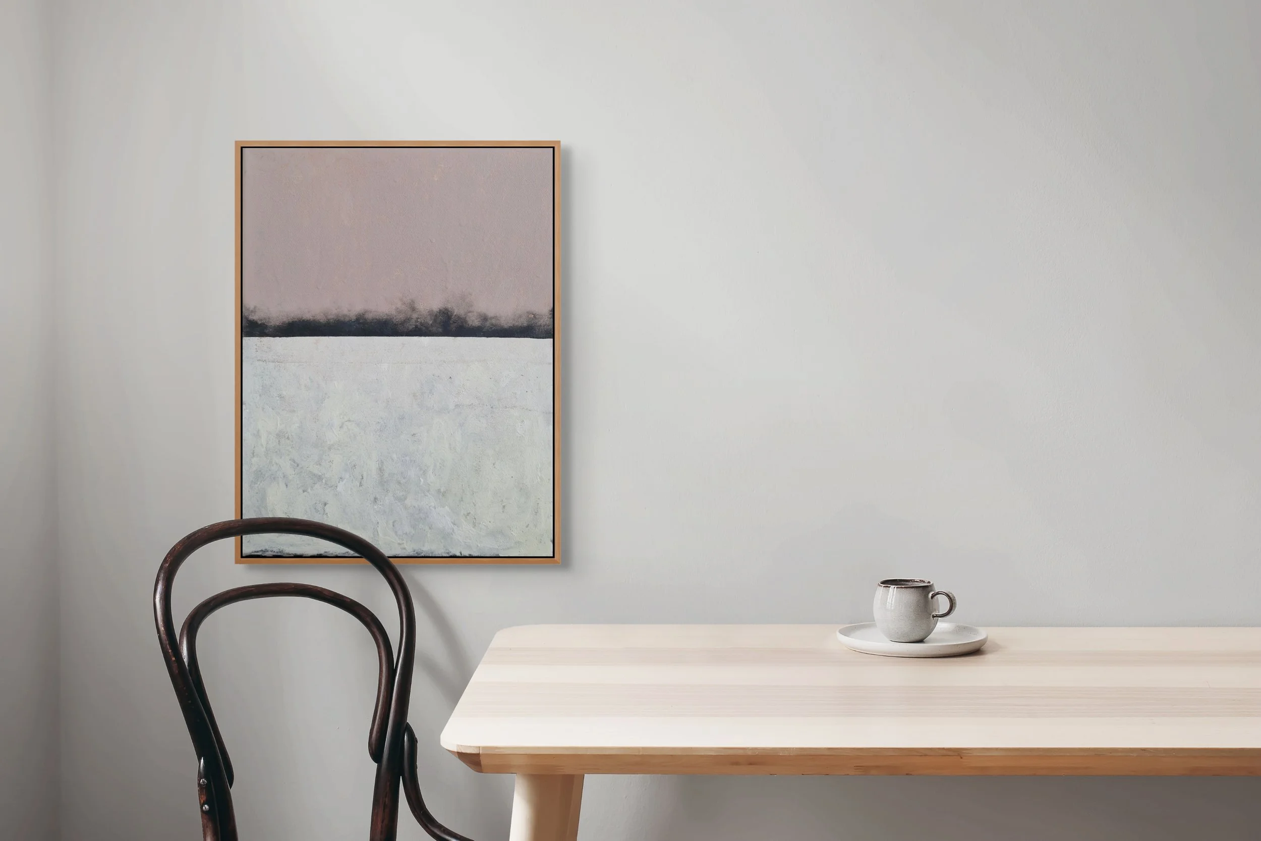 Minimalist interior with a light wooden table, a ceramic mug on a plate, two dark wooden chairs, and an abstract painting on a white wall.