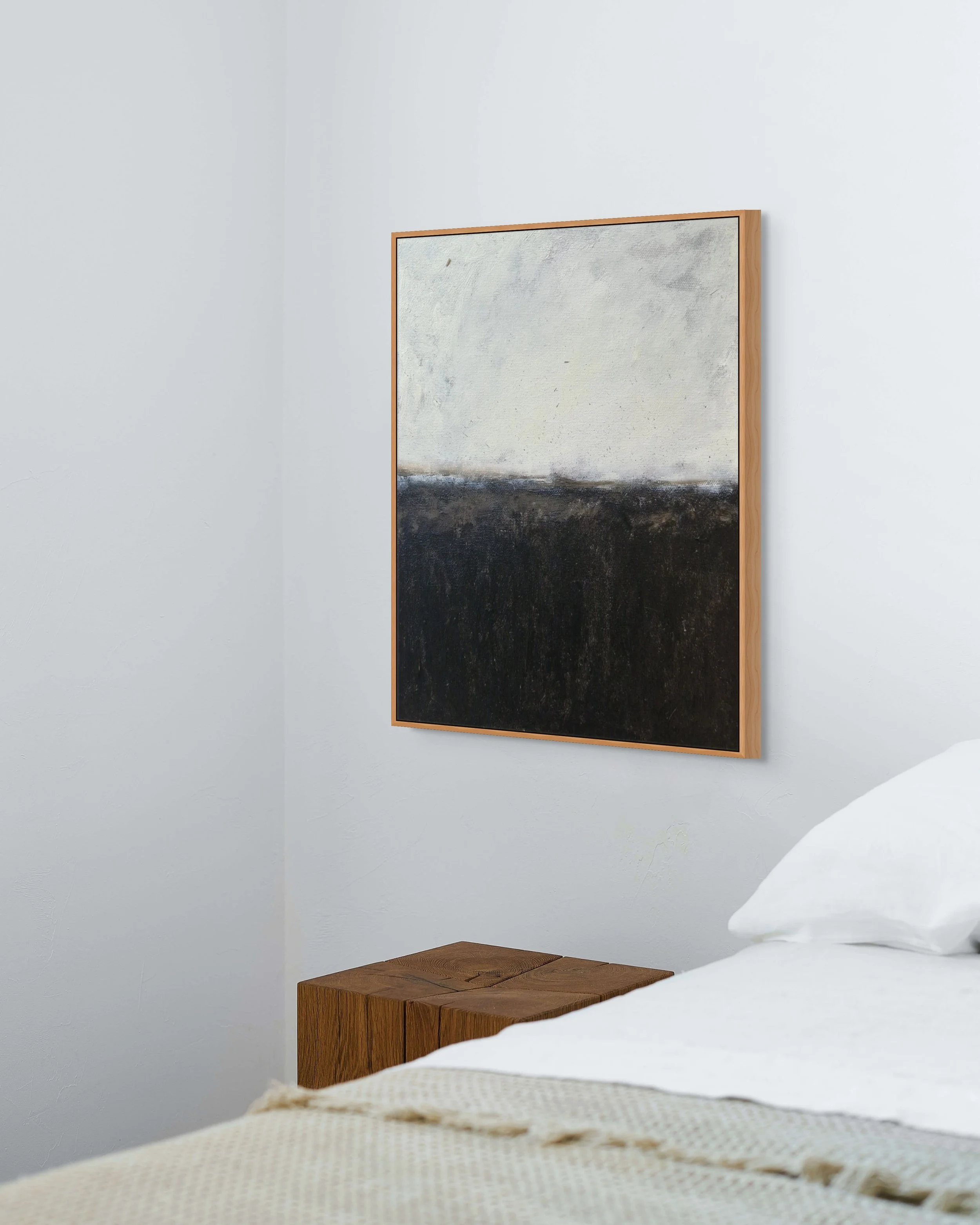 A bedroom with a white wall, a wooden nightstand, and an abstract painting with a light upper half and dark lower half framed in wood.