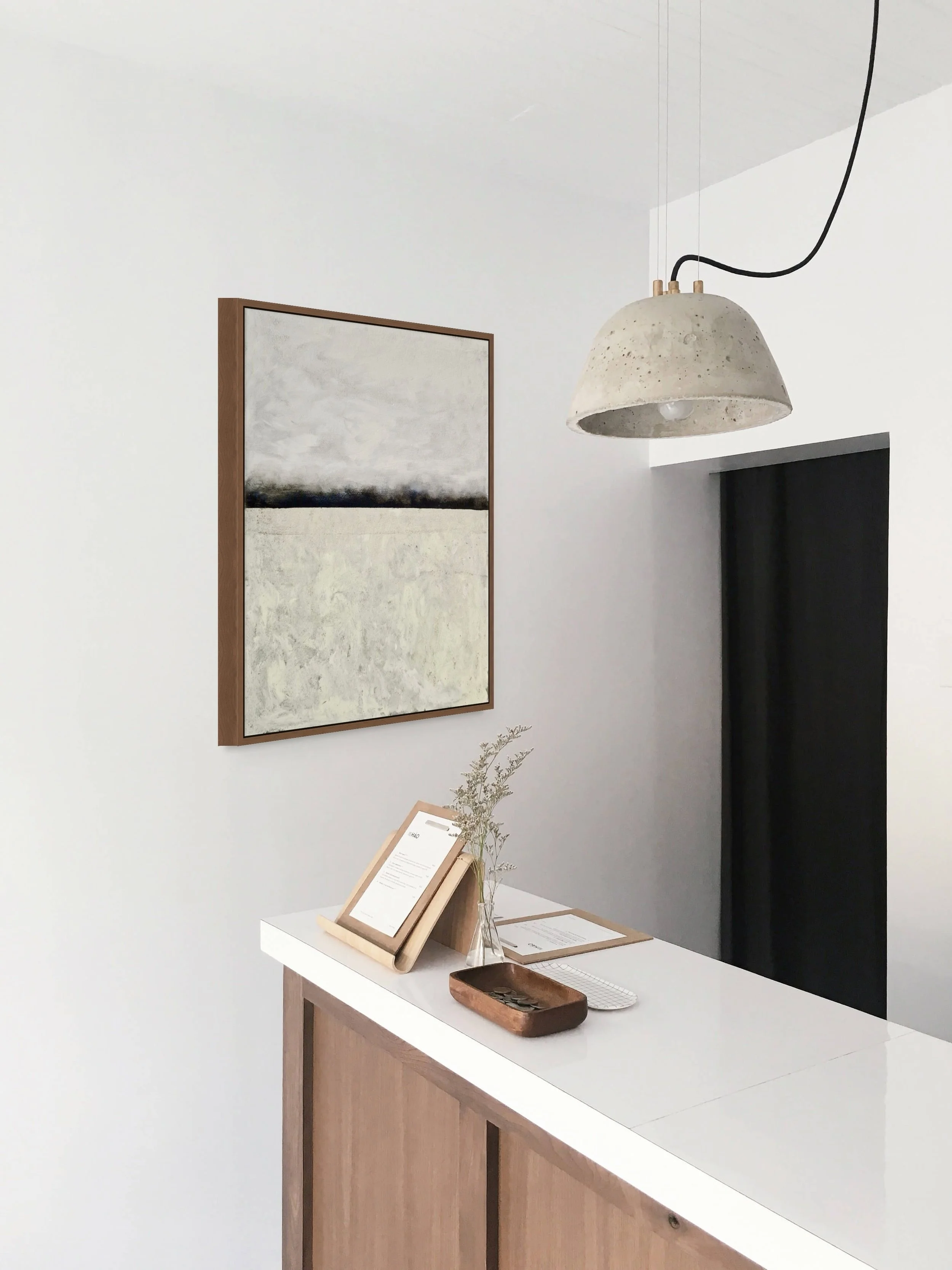 Minimalist reception desk with framed menu, glass vase with dried plant, wooden tray, and framed papers on a white surface, with abstract black-and-white painting on the wall and a concrete pendant light hanging above.
