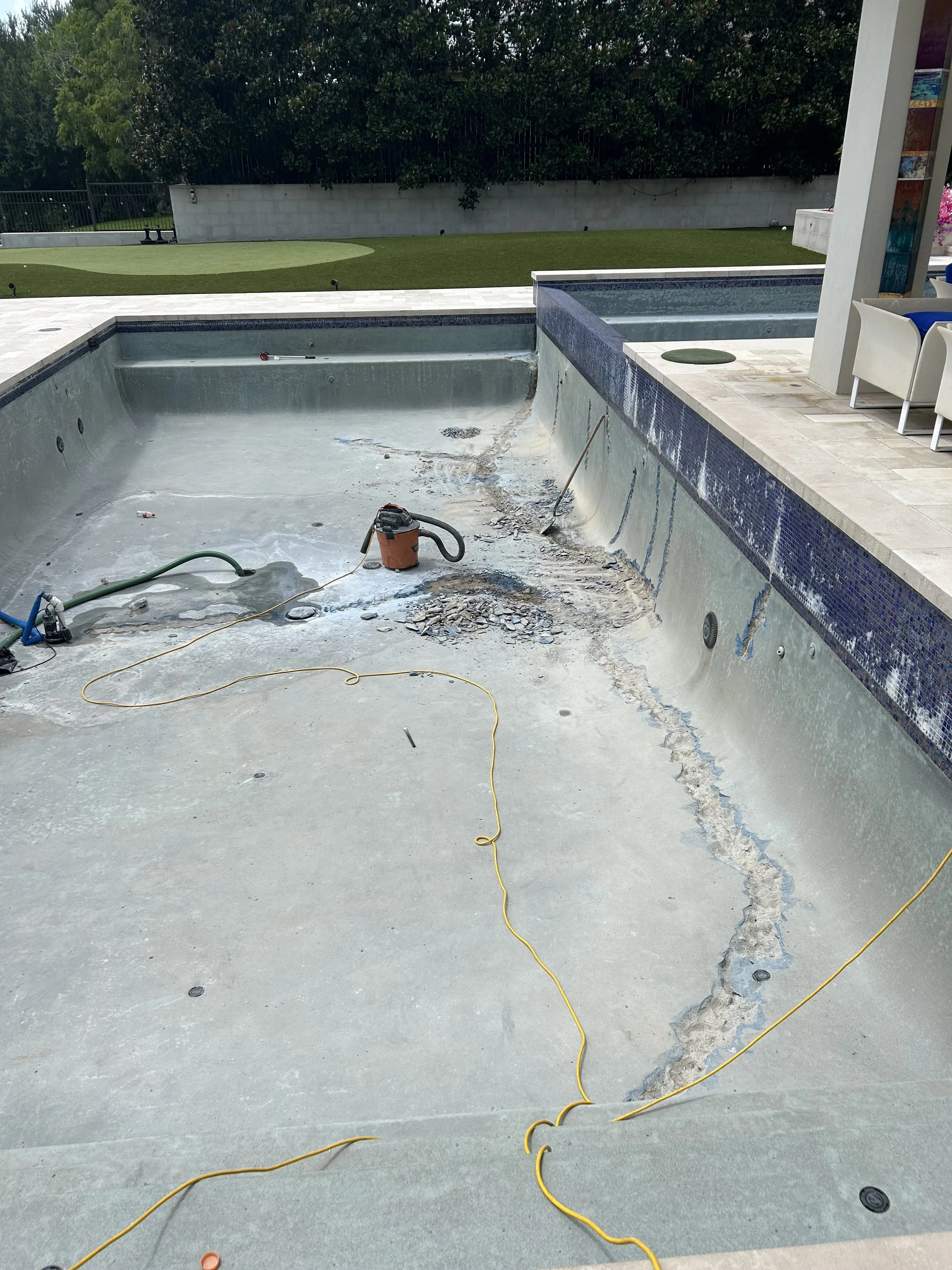 An empty swimming pool undergoing renovation, with exposed concrete and construction equipment such as a vacuum cleaner and hoses. A garden with neatly trimmed grass is visible in the background.