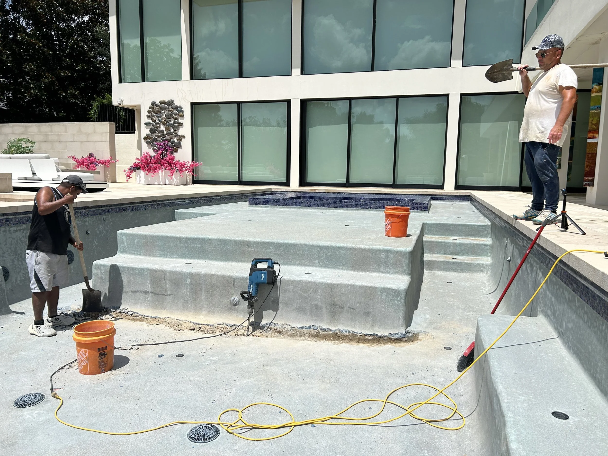 Two workers standing in an empty swimming pool under renovation, with tools and equipment. Modern building with large windows visible in the background.