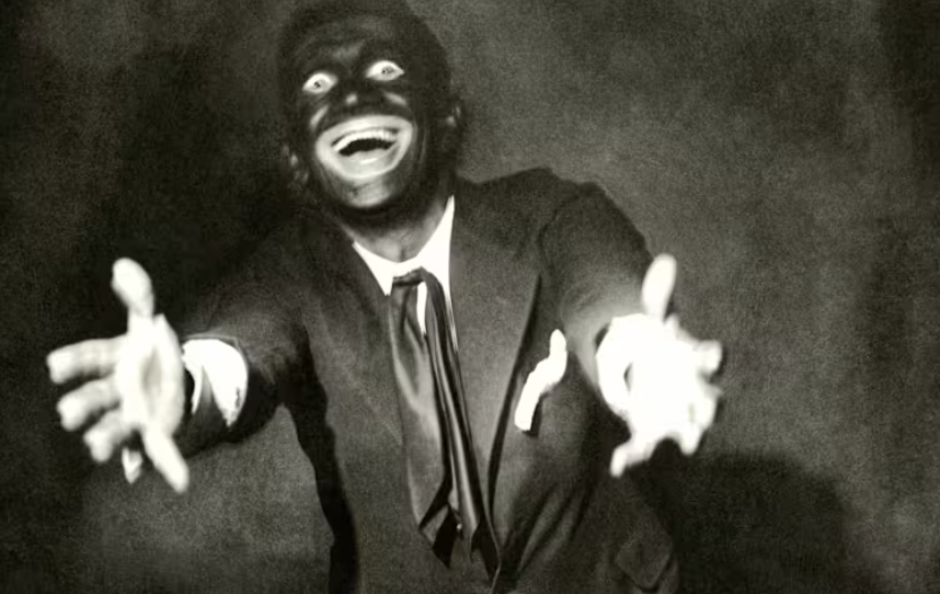 Al Jolson, a famous minstrel performer in a suit with a tie, smiling widely with eyes wide open, reaching out with both hands.