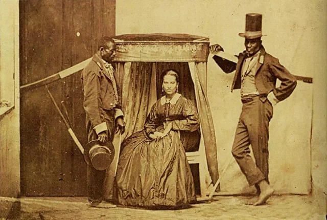 An old photograph showing a seated woman in the center, flanked by two enslaved men in Brazil.