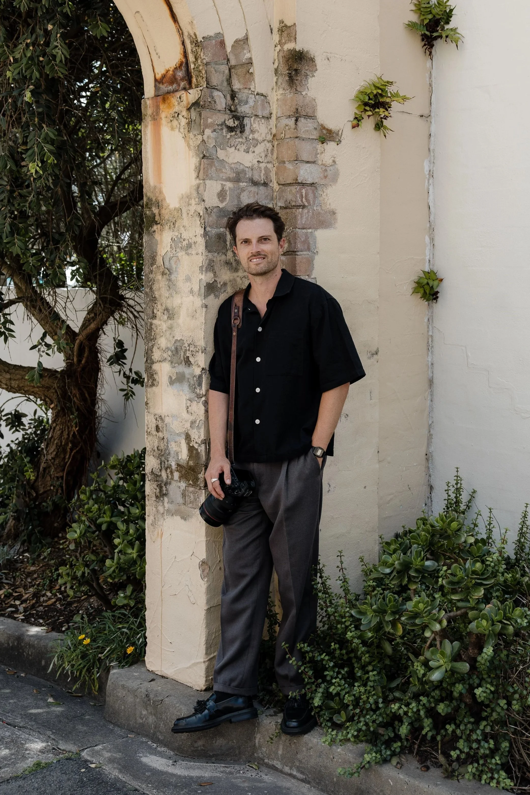 A young man with dark, wavy hair and a light beard standing outdoors against a weathered, beige brick and stucco wall. He is wearing a black short-sleeved button-up shirt, gray trousers, black dress shoes, and a watch. He is holding a camera in his right hand and has a brown strap over his shoulder. There are green bushes at his feet and a leafy tree on his left side.