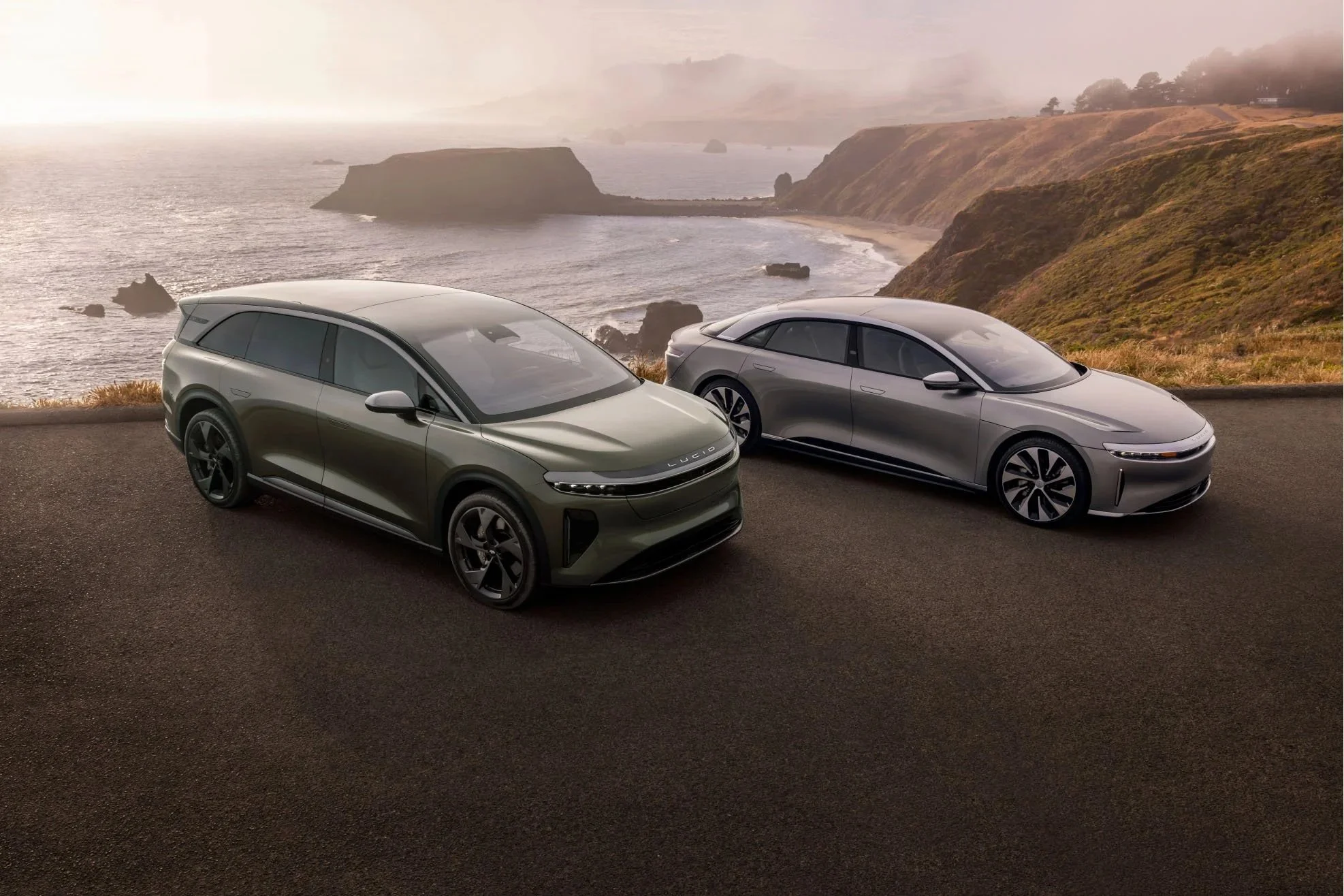 Zwei moderne Elektroautos stehen an einer Landstraße mit Blick auf eine Küstenlandschaft bei Sonnenaufgang oder Sonnenuntergang, mit Felsen im Meer und hügeligem Gelände im Hintergrund.