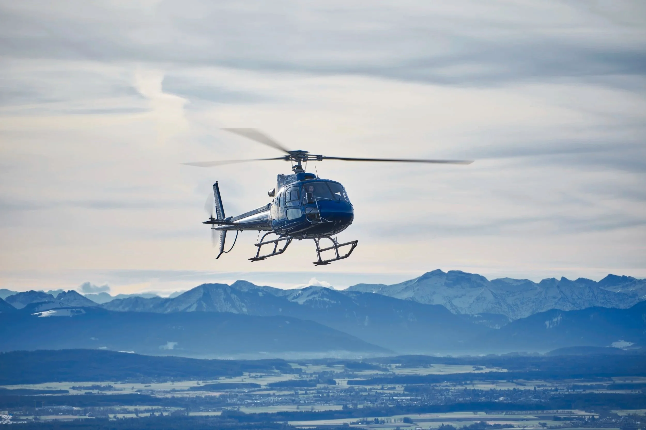 Ein blauer Helikopter fliegt vor einer Bergkulisse durch den Himmel.