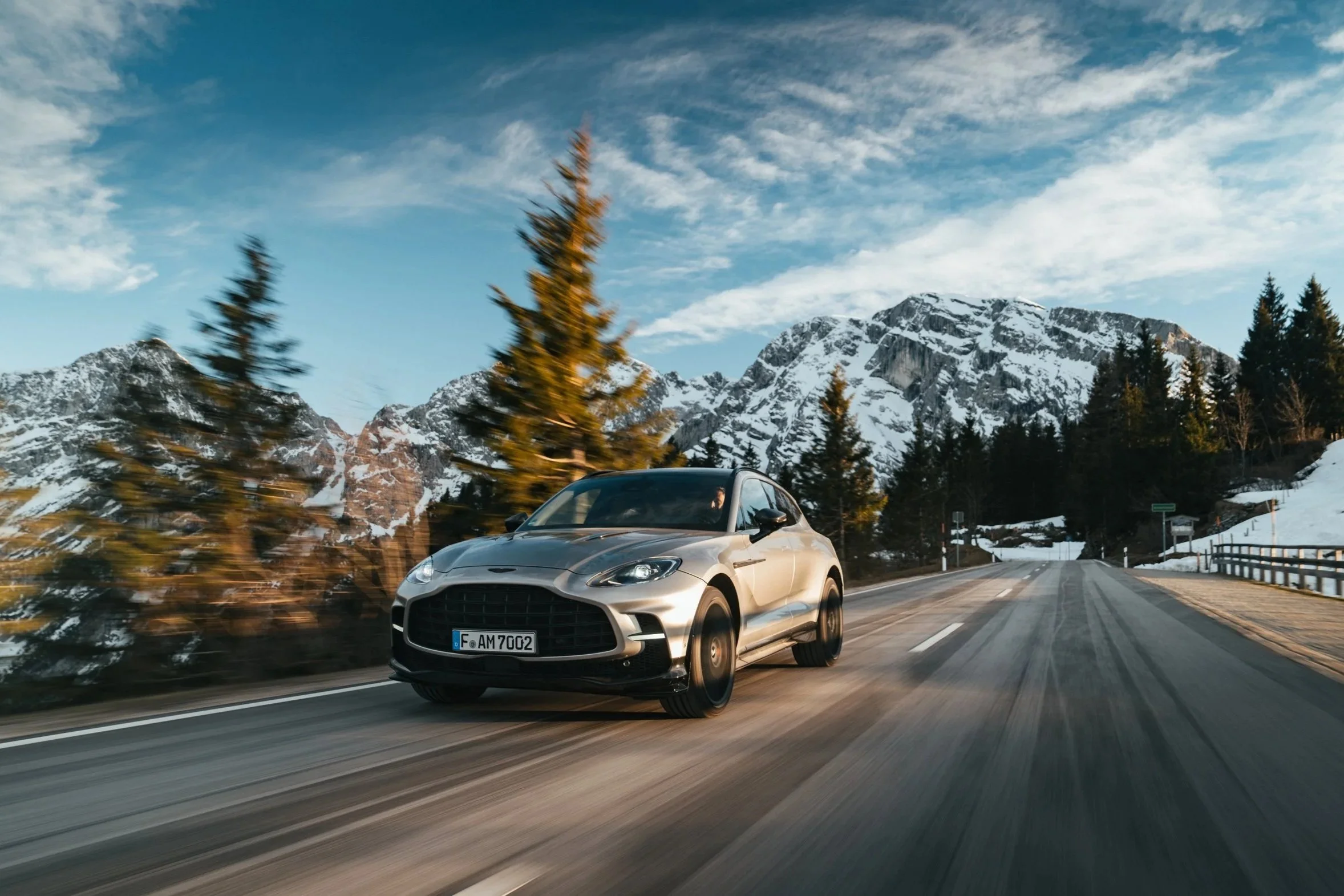 Ein silberner Sportwagen fährt auf einer kurvigen Straße in den Bergen, umgeben von Tannenbäumen und schneebedeckten Bergen unter einem blauen Himmel mit Wolken.
