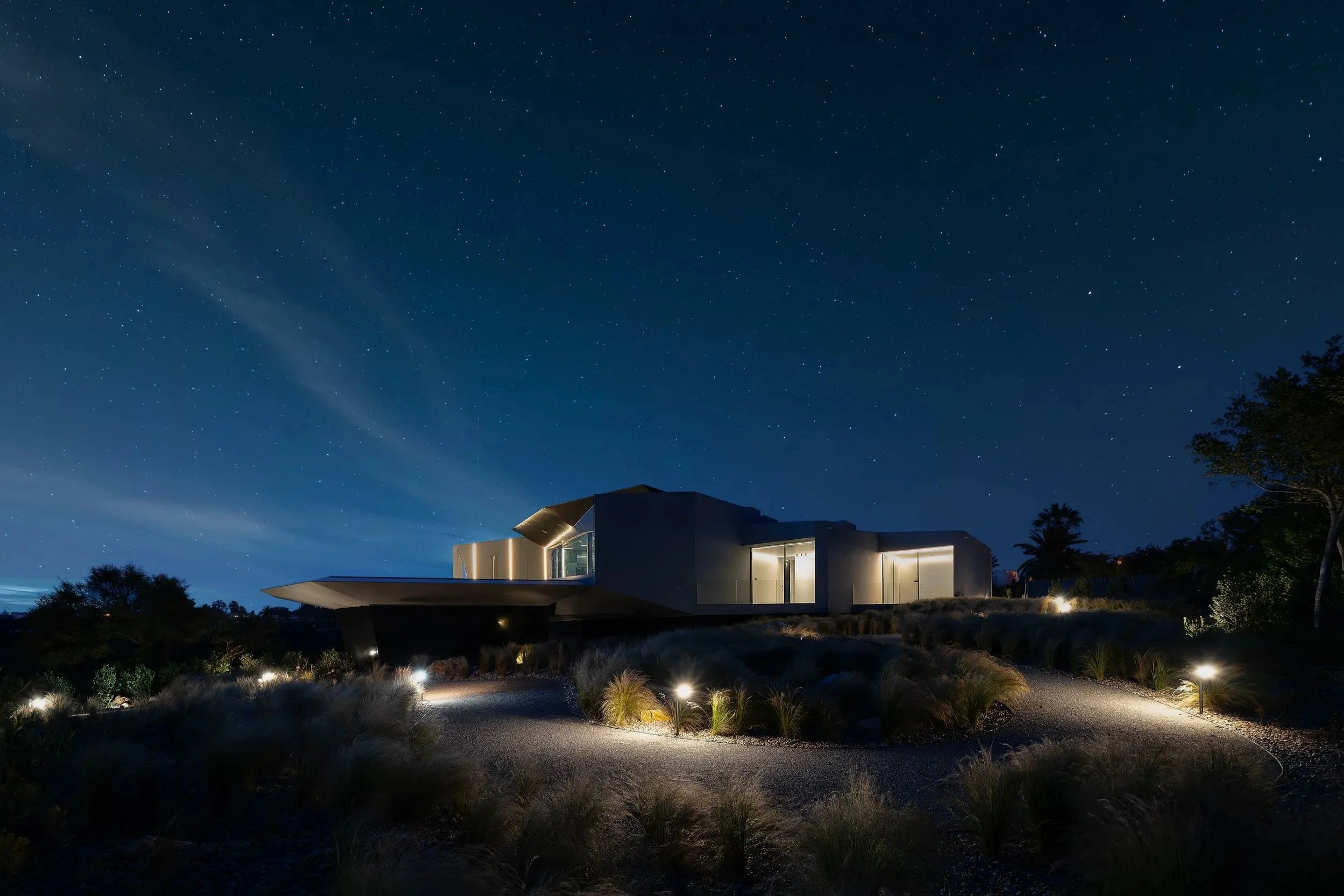 Moderne weiße Villa bei Nacht mit beleuchteten Fenstern und Gartenbeleuchtung, umgeben von Rabatten und Büschen, unter einem klaren Sternenhimmel.