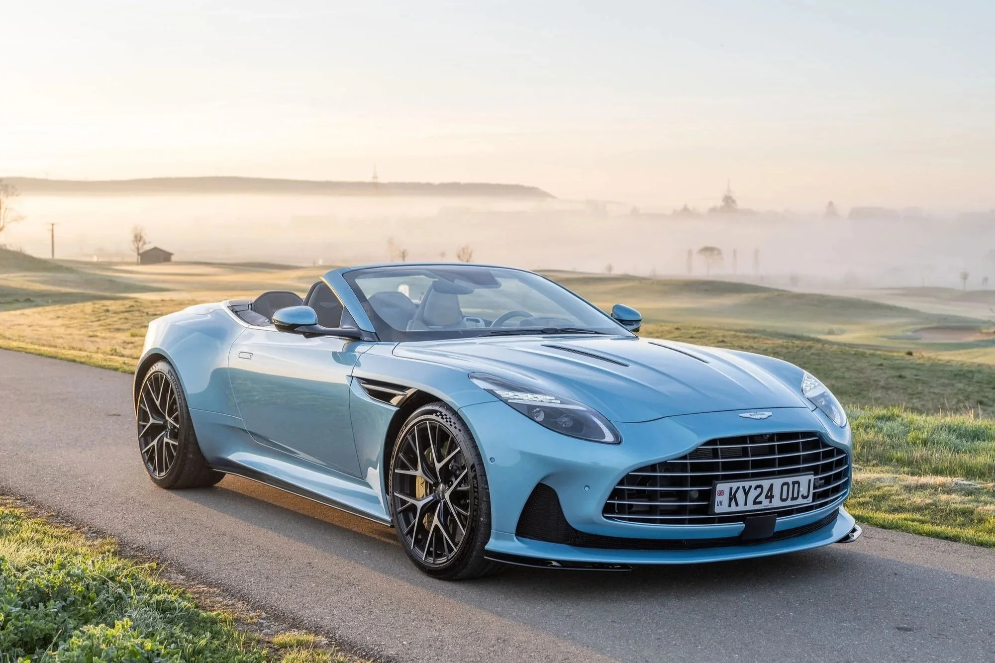 Blauer Sportwagen, vermutlich ein Aston Martin, vor einer nebligen Landschaft im Morgengrauen, mit Hügeln und Bäumen im Hintergrund.