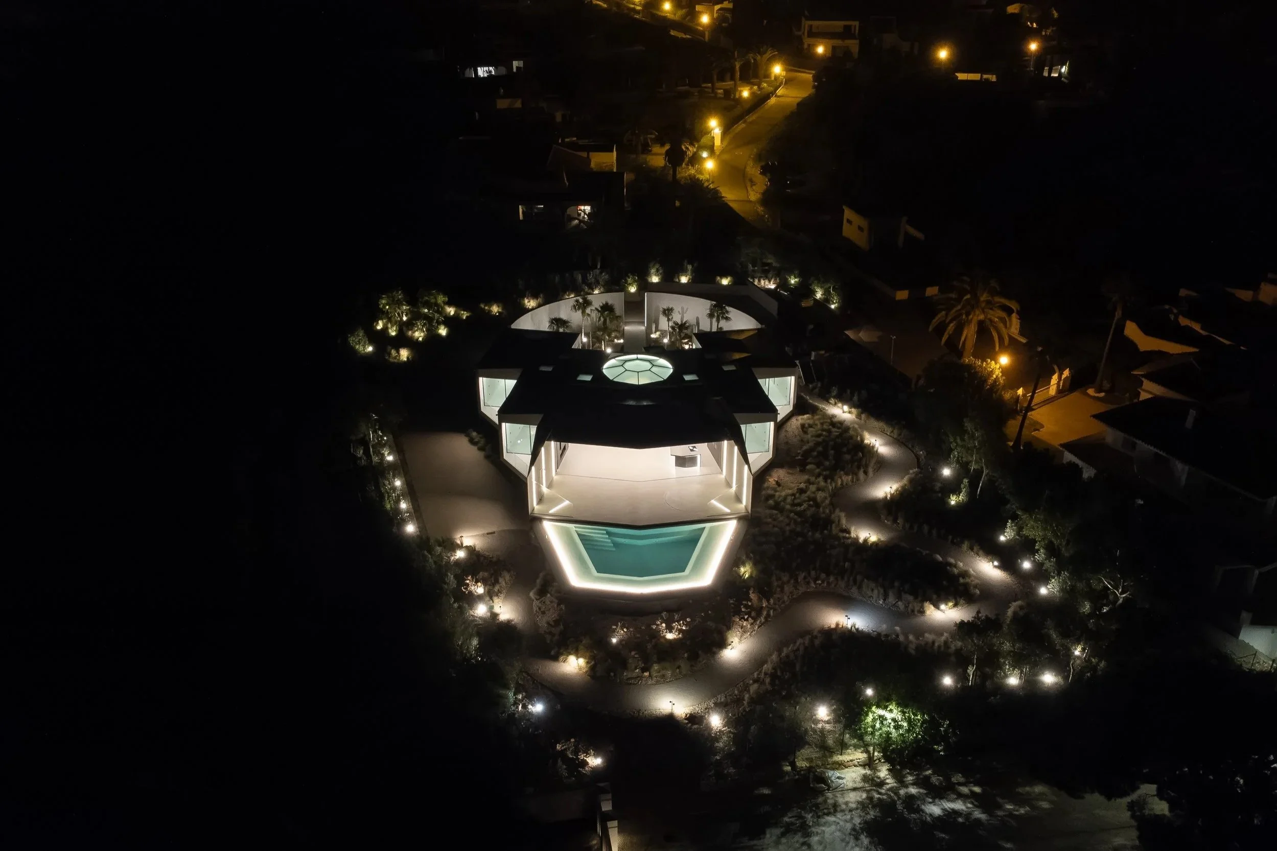 Ein moderner, beleuchteter Wohnbereich mit Pool vor einem schwarzen Haus, umgeben von Bäumen und einer Straße mit Straßenlaternen bei Nacht.