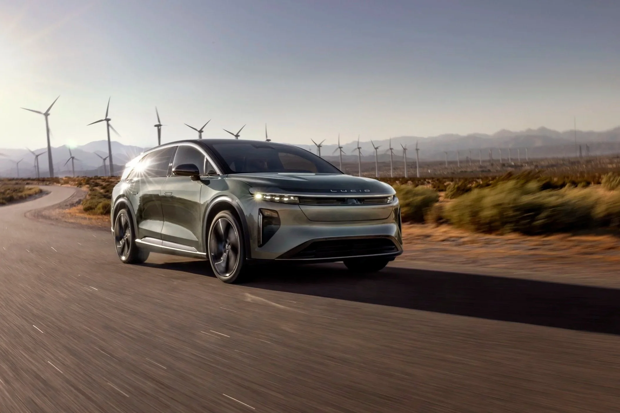 Ein futurisches Elektroauto fährt auf einer Landstraße mit Windkraftanlagen im Hintergrund unter klarem Himmel.