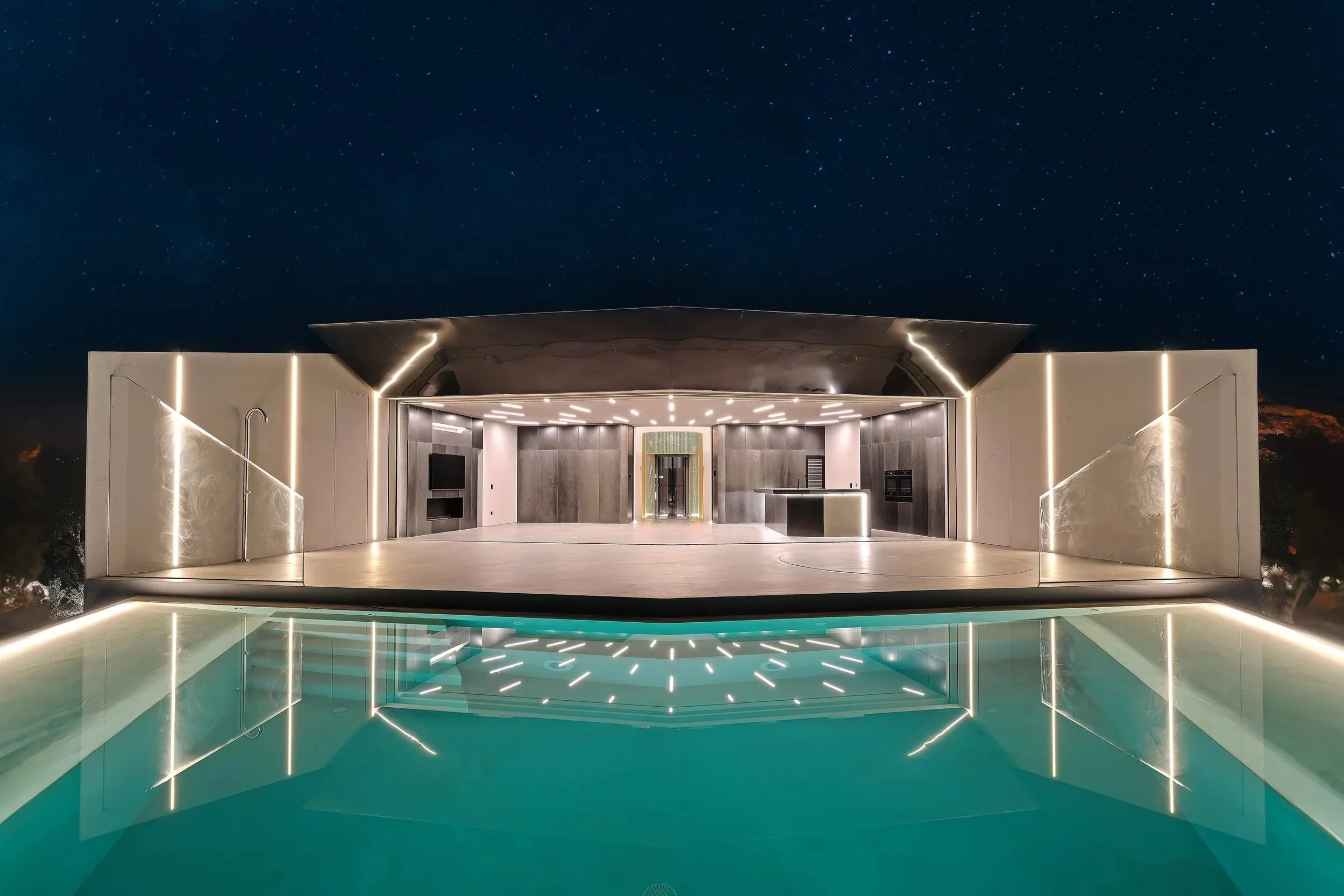 Moderner Gebäudekomplex mit beleuchteter Außenfassade über einem reflektierenden Pool bei Nacht, Sternenhimmel im Hintergrund.