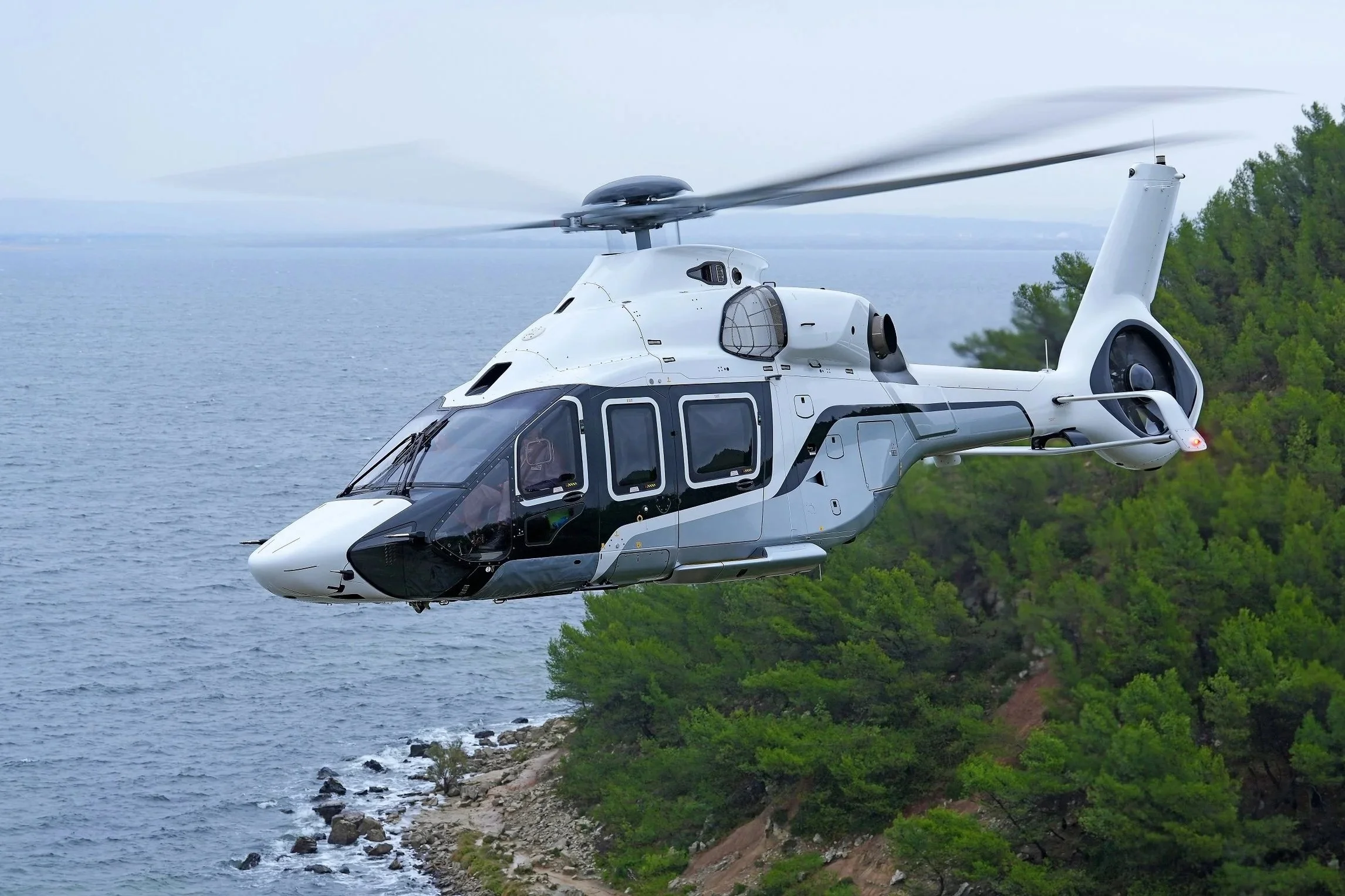 Ein modernes Helikopter fliegt über das Wasser mit grünen Bäumen im Hintergrund.