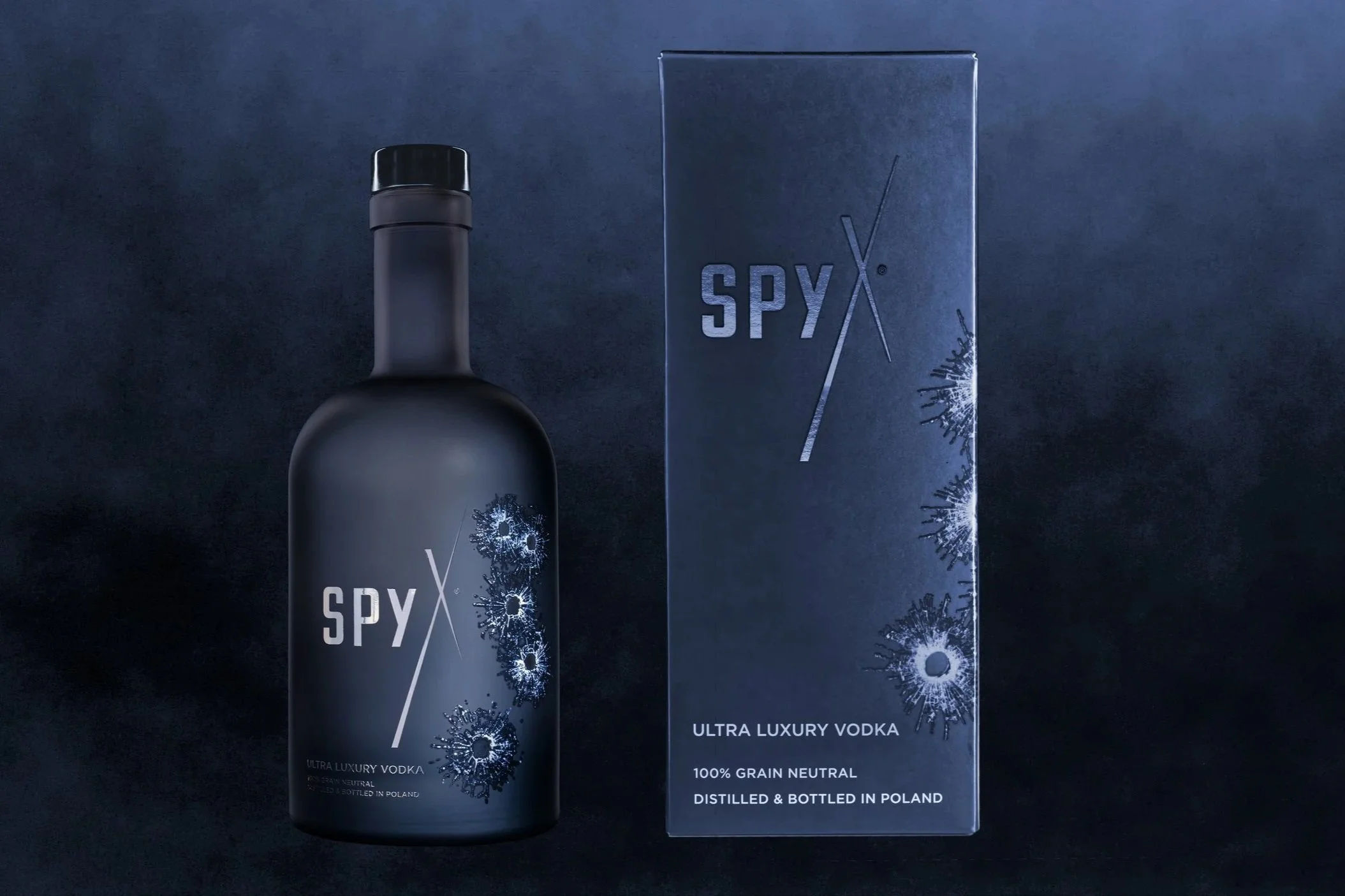 Eine Flasche und ein Verpackungskarton von Spy X Ultra Luxury Wodka auf dunklem Hintergrund. Die Flasche ist matt schwarz mit einem schwarzen Verschluss und einem Dekor aus blauen Explosionen. Das Etikett auf der Flasche weist auf 100% Grain Neutral,