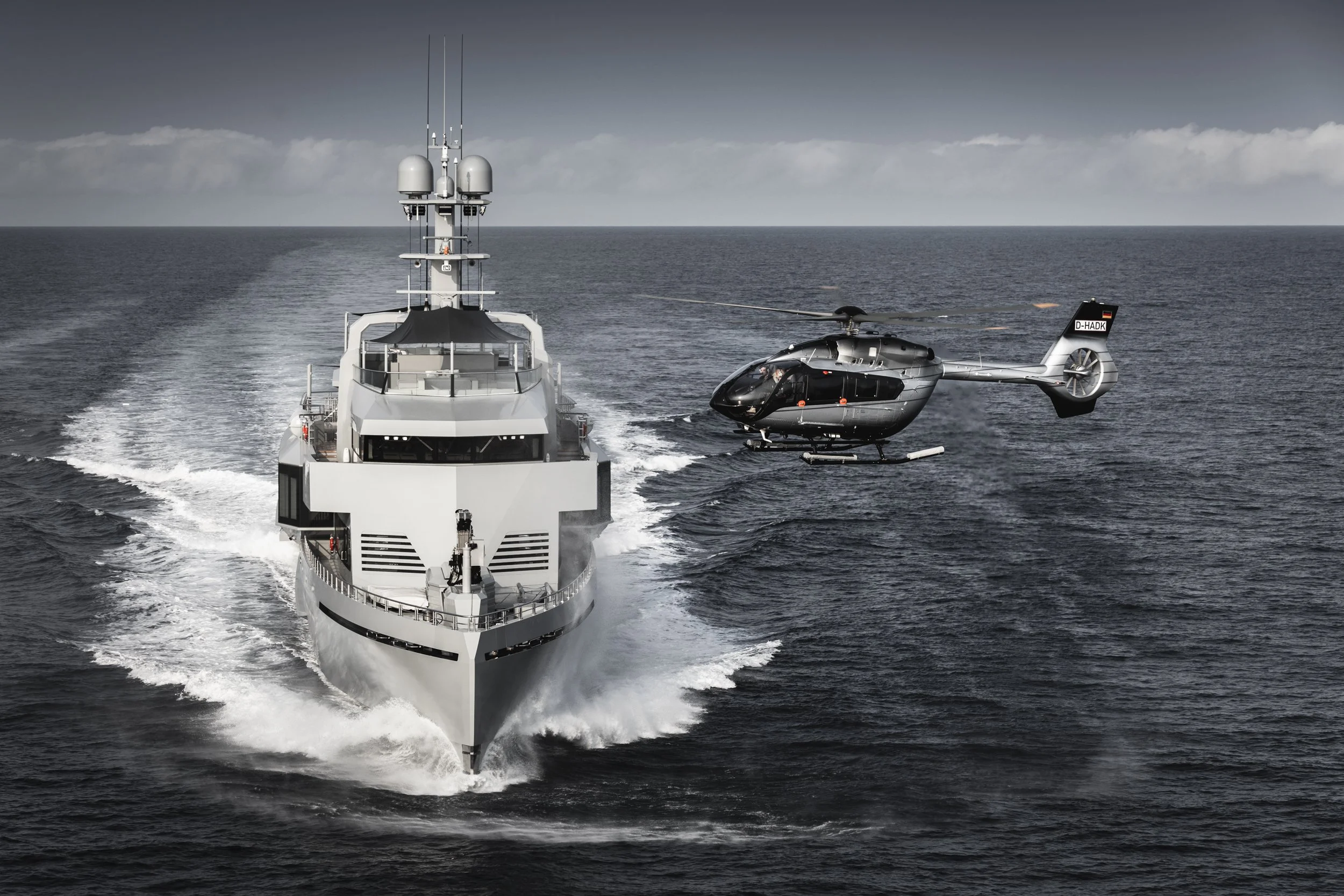 Eine weiße Yacht fährt auf dem Meer, im Hintergrund ist ein Hubschrauber über dem Wasser unterwegs.