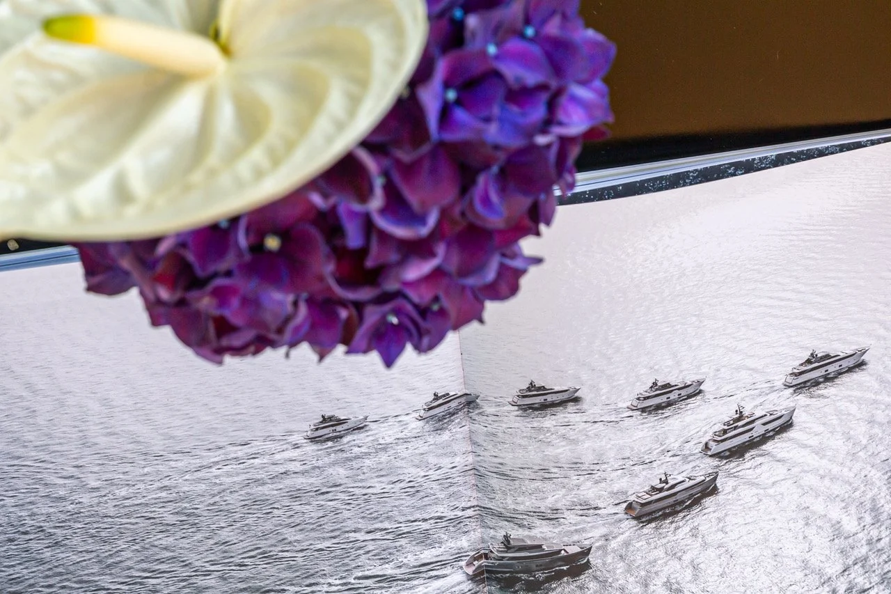 Blick auf das Wasser mit mehreren vorbeifahrenden weißen Yachten, im Vordergrund eine Vase mit lilafarbenen Hortensien und eine weiße Blume.