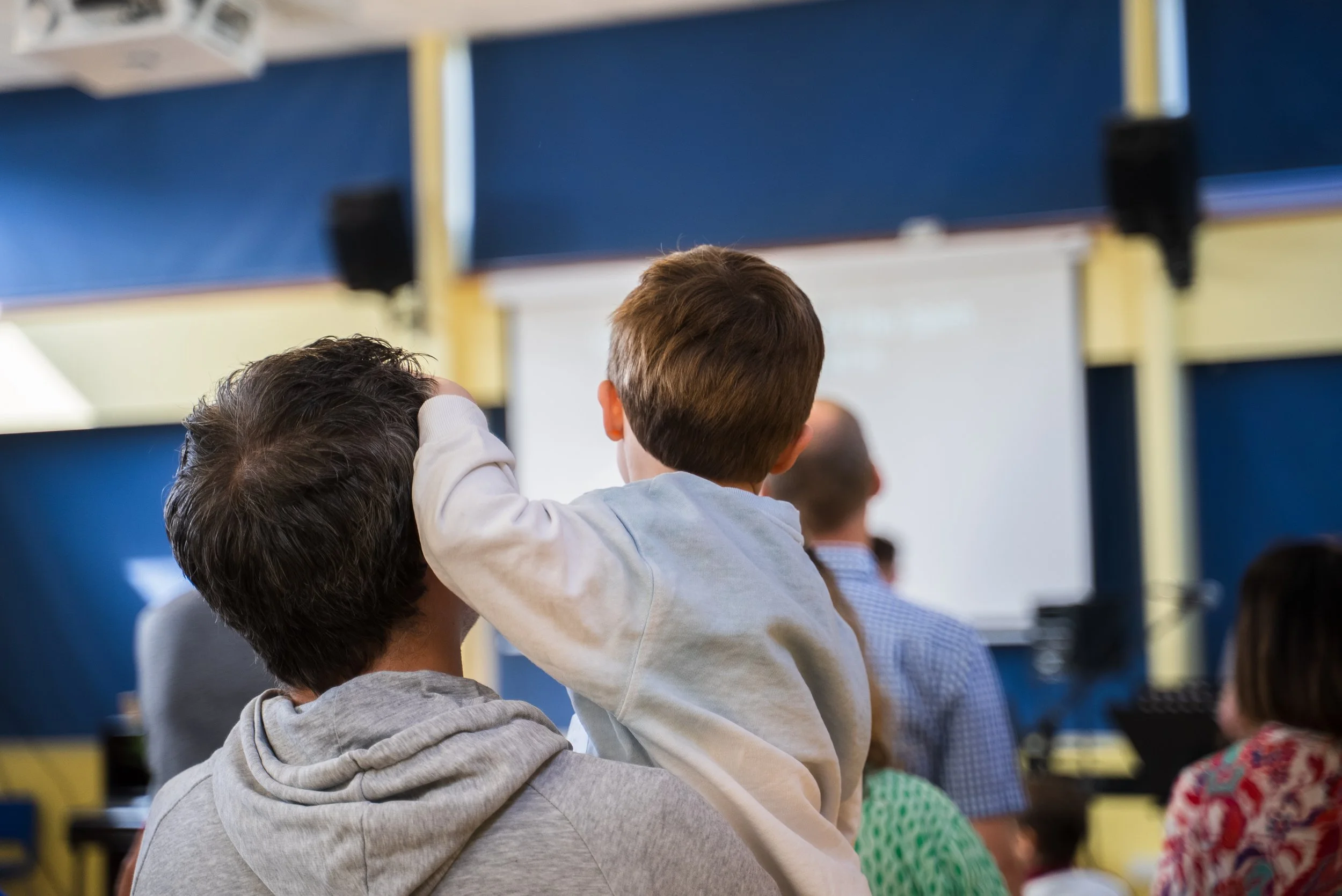 A young boy sitting on an adult's shoulder in a classroom or auditorium, looking at a whiteboard or screen at the front of the room, surrounded by other seated people.