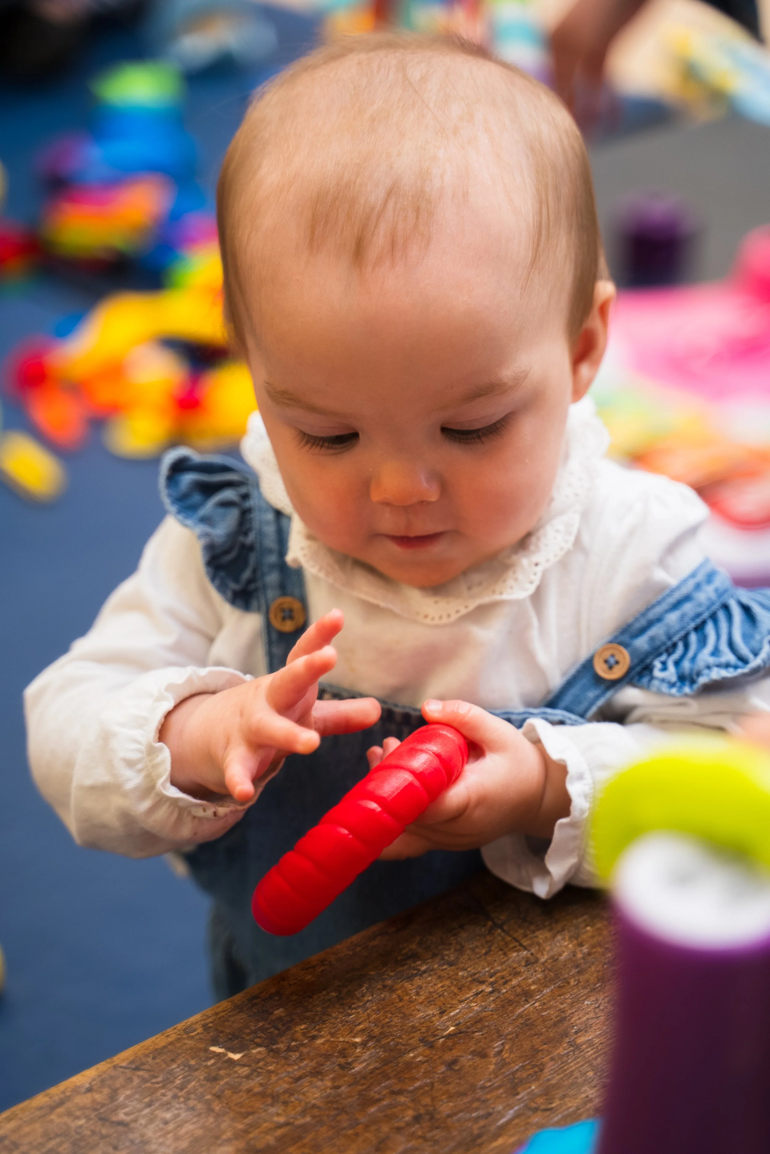 A young child with light hair and a white top with ruffled sleeves holding a red toy and looking at it carefully in a colorful play area.