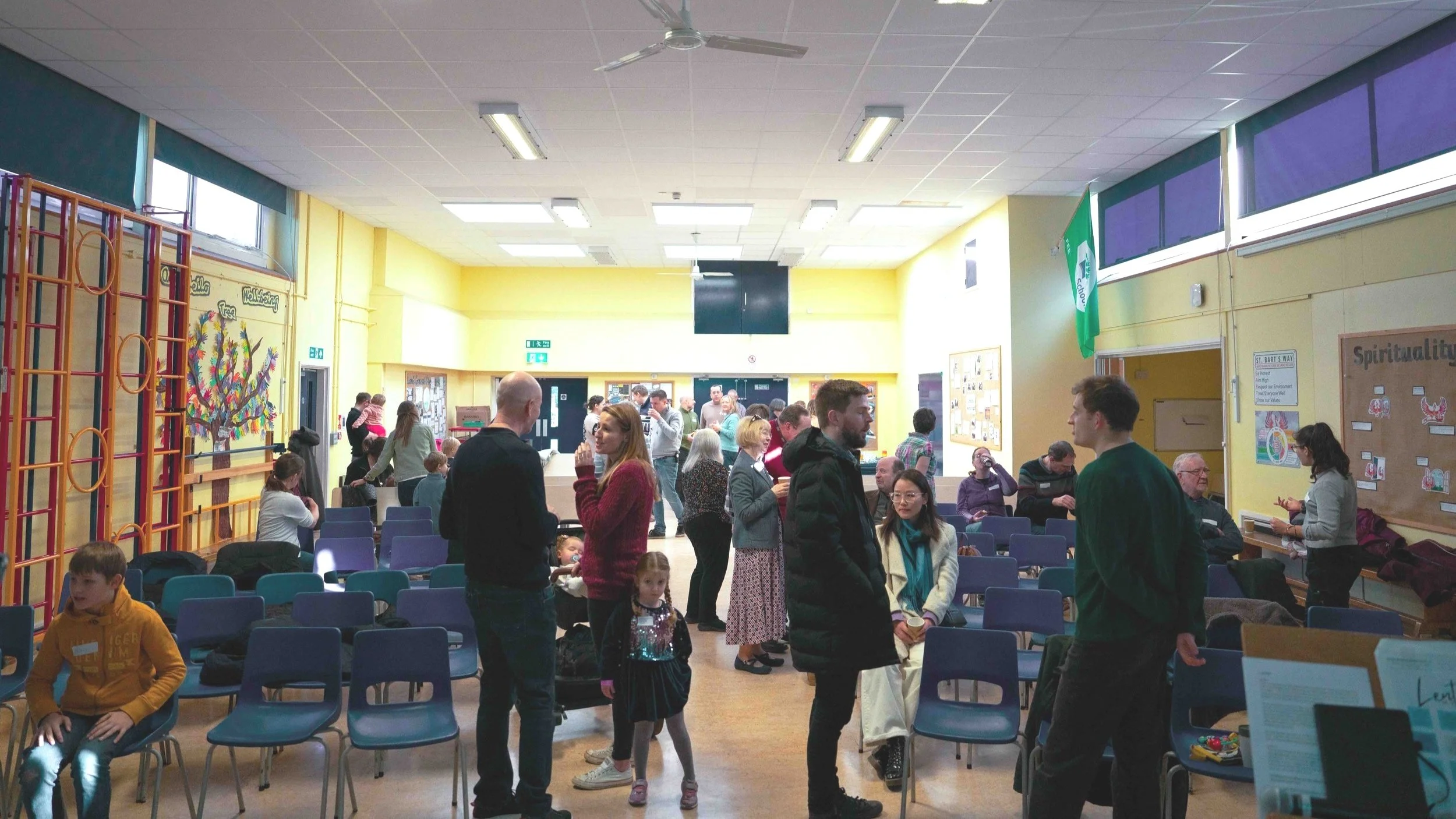 A large room filled with people, including children and adults, some seated and some standing, engaging in conversations. The room has yellow walls, blue chairs, and bulletin boards on the walls. There are windows at the top, and the lighting is bright and well-lit.