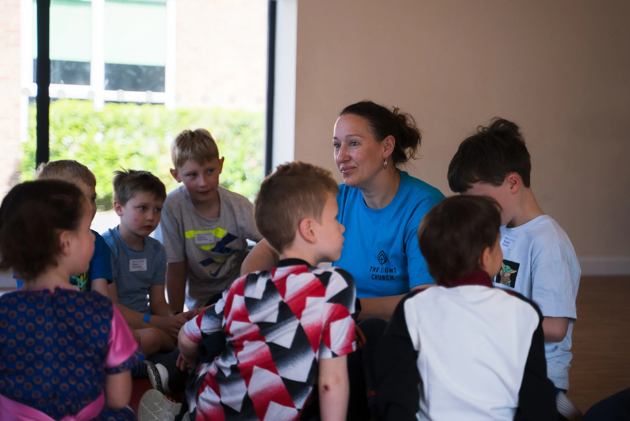 A woman with dark hair, wearing a blue shirt, talking to a group of children indoors near a window.