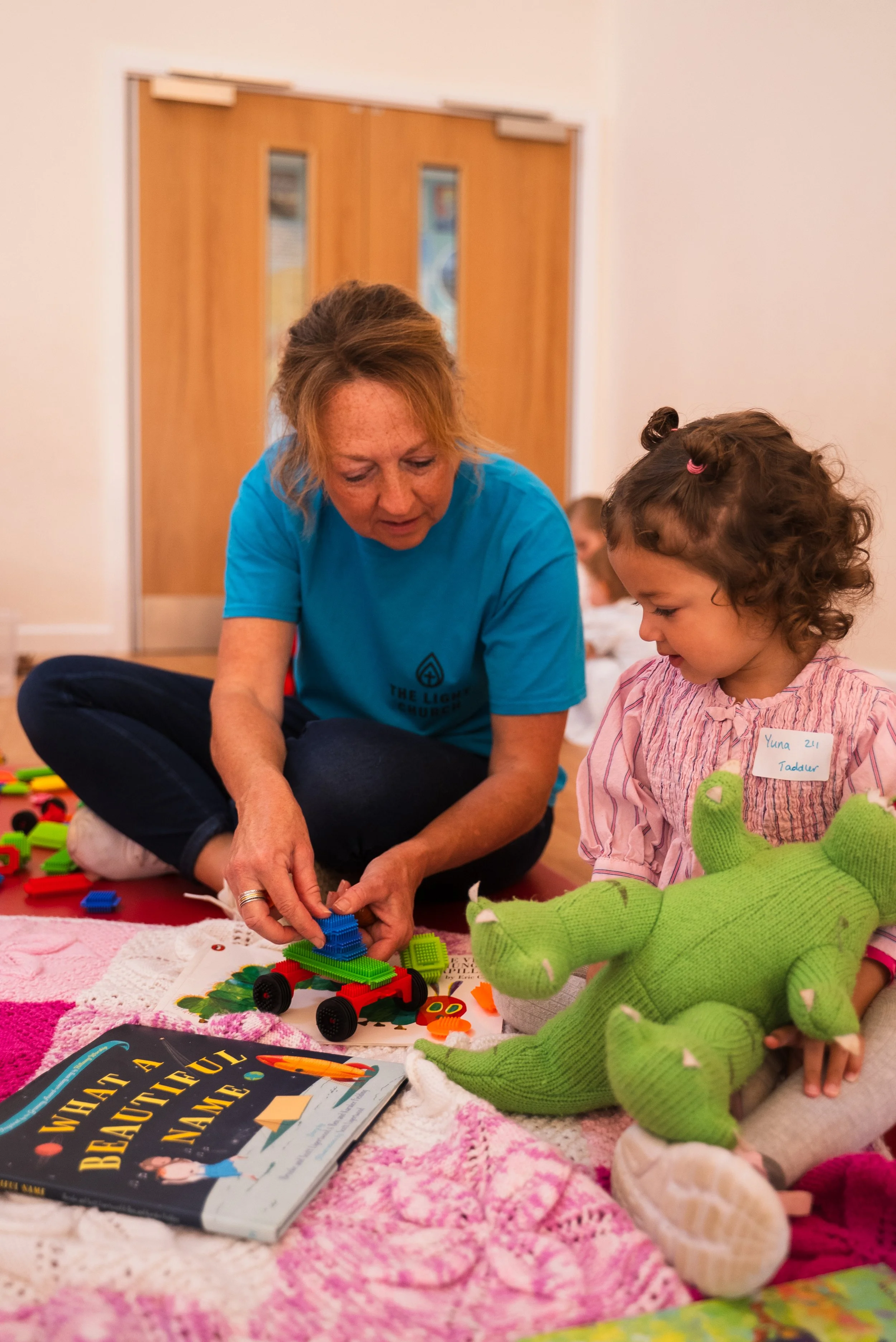 An adult woman and a young girl sitting on the floor playing with colorful plastic building blocks, with a green stuffed toy and a children’s book nearby.